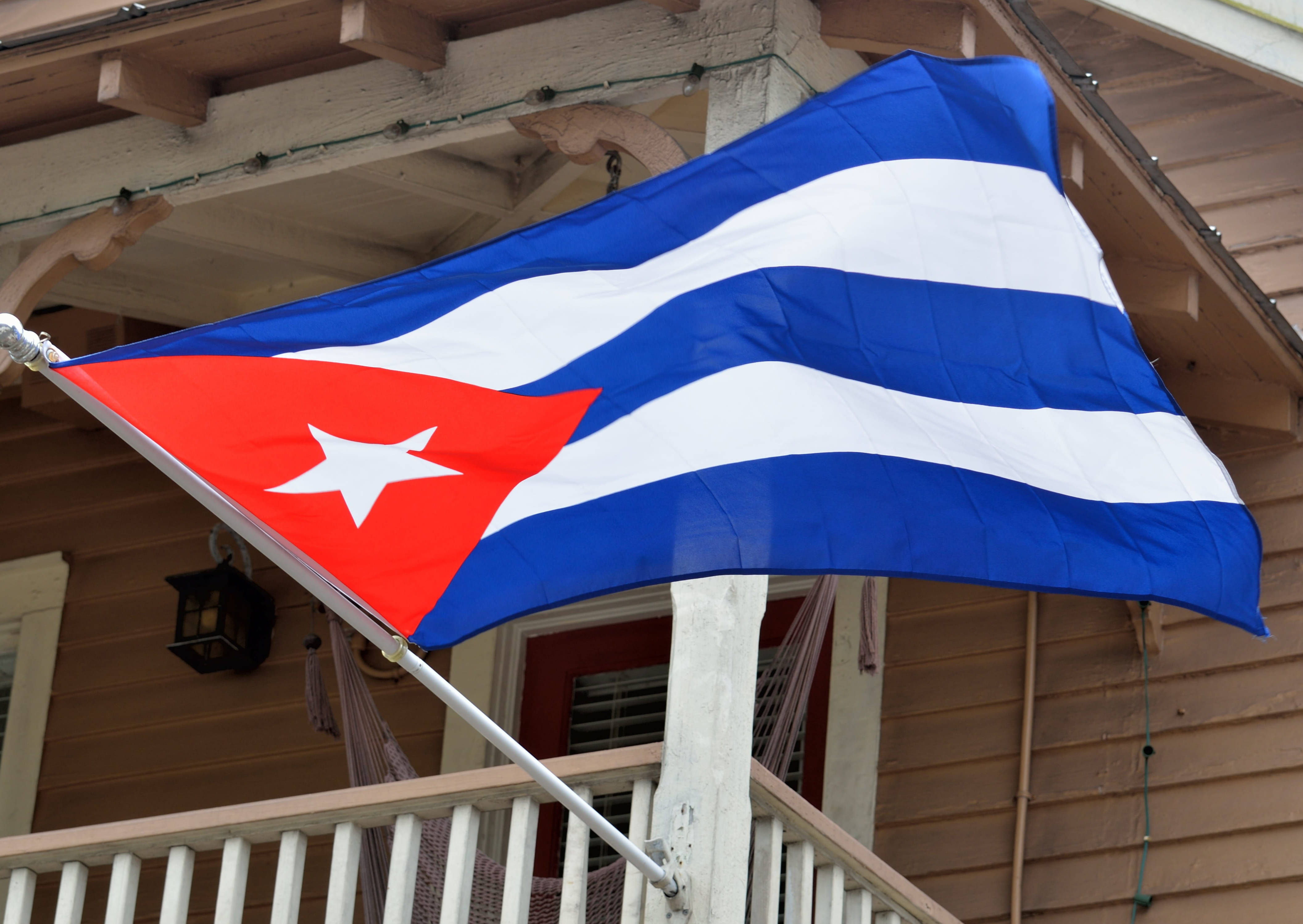 Cuban Flag 桌布 壁紙 — 帶有紅星和藍白條紋的古巴國旗在木質牆板和串燈襯托下，在門廊上大膽飄揚。