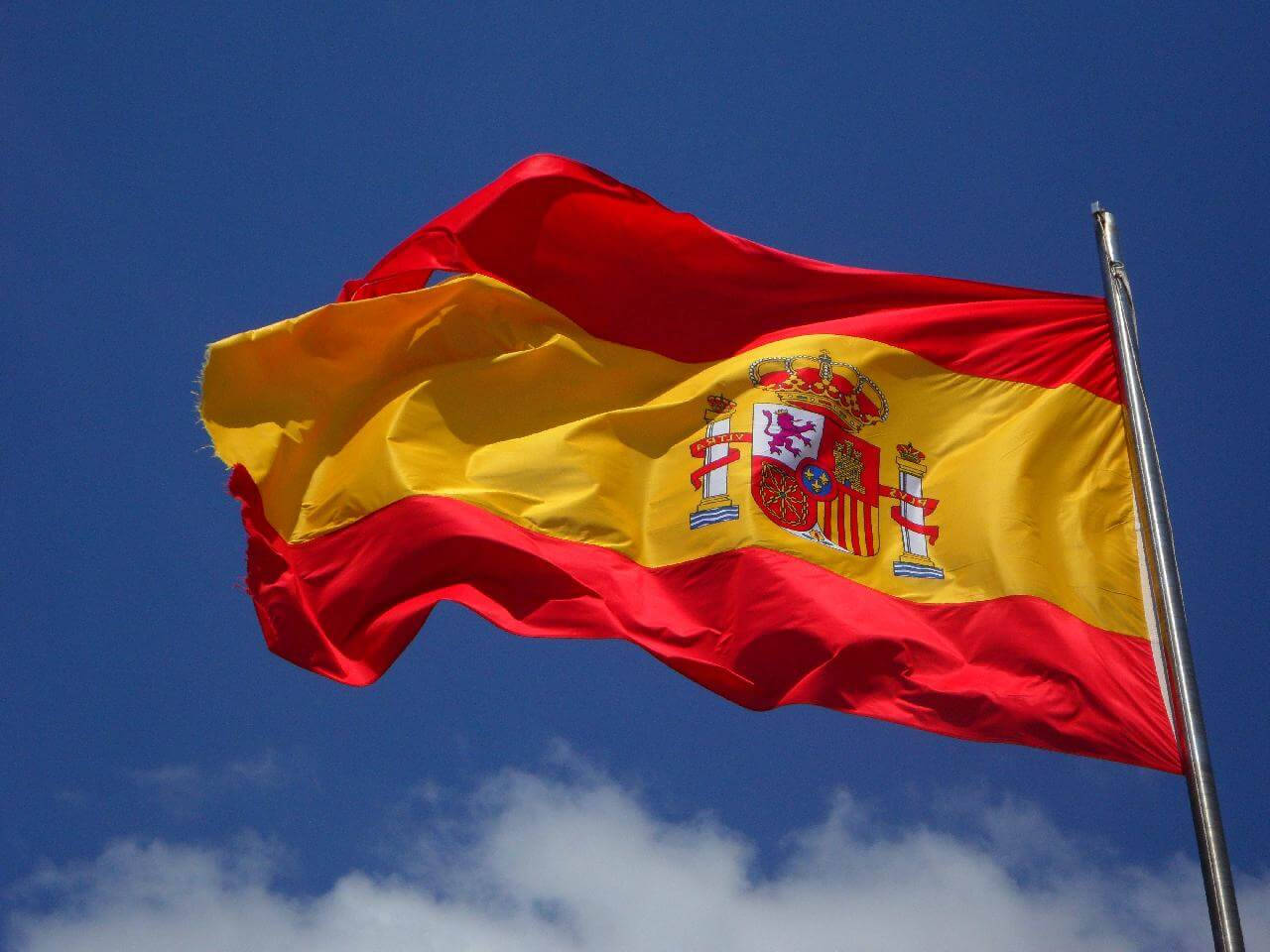 Spain Flag 桌布 壁紙 — 帶有紅色和黃色條紋及國徽的西班牙國旗在藍天白雲中自豪地飄揚。