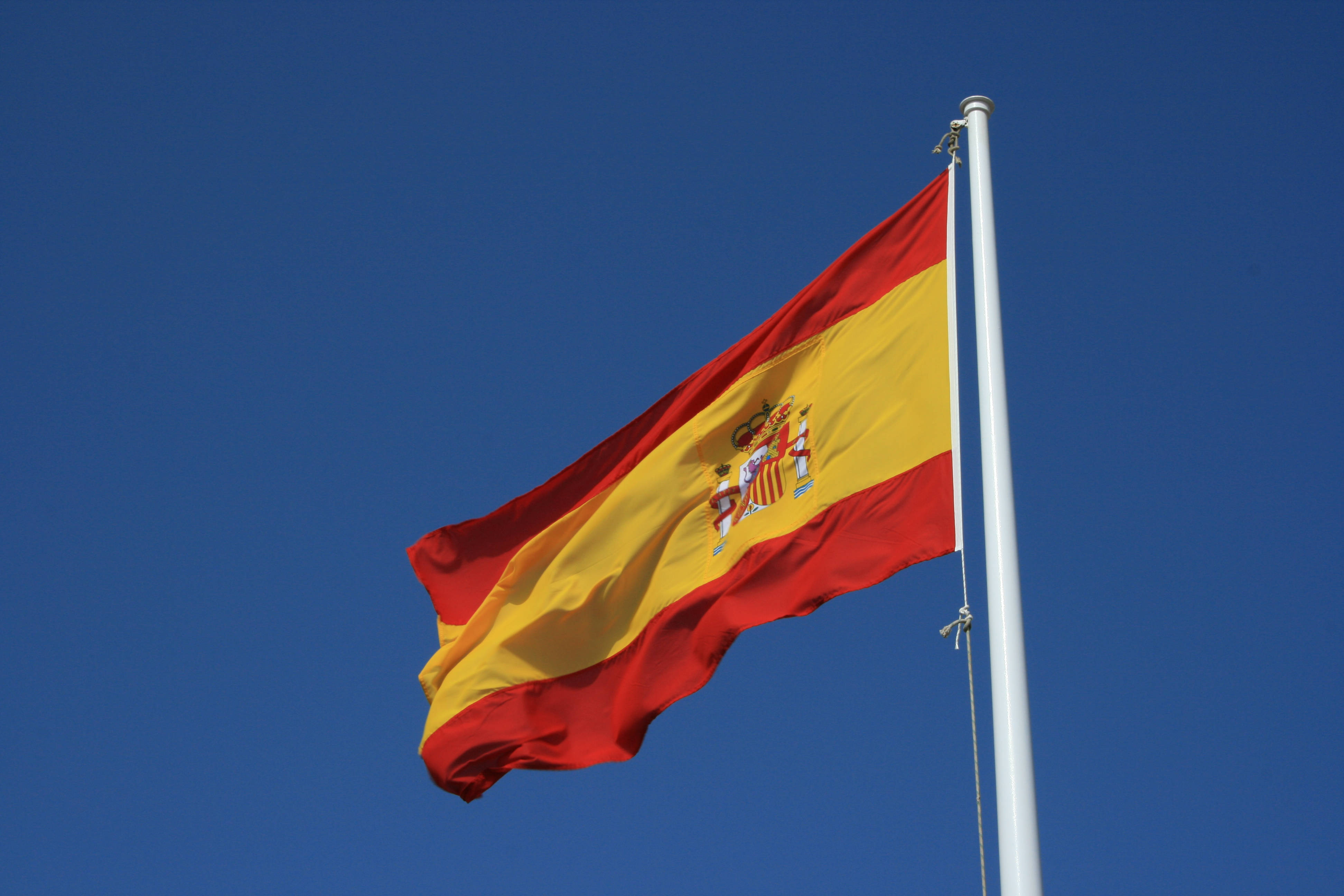 Spain Flag 桌布 壁紙 — 帶有紅黃條紋和國徽的西班牙國旗在晴朗的藍天下自豪地飄揚。