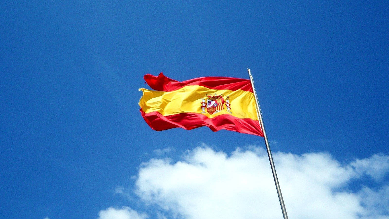 Spain Flag 桌布 壁紙 — 帶有紅黃條紋和國徽的西班牙國旗在藍天白雲中自豪地飄揚。