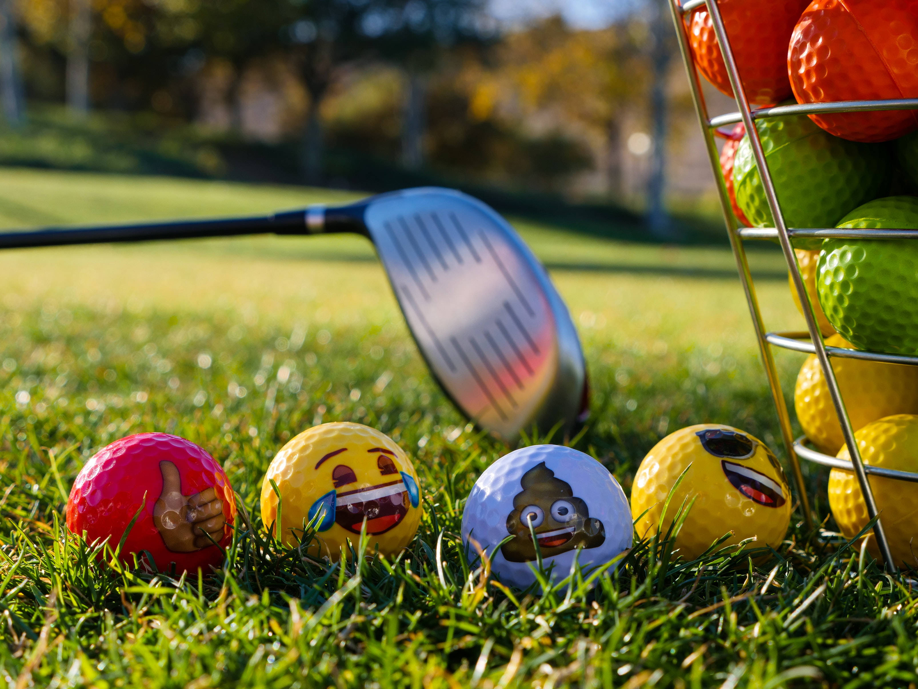 4K Golf 桌布 壁紙 — 帶有表情符號臉的高爾夫球放在草地上，旁邊有球桿和色彩繽紛的球架，在明亮的陽光下。