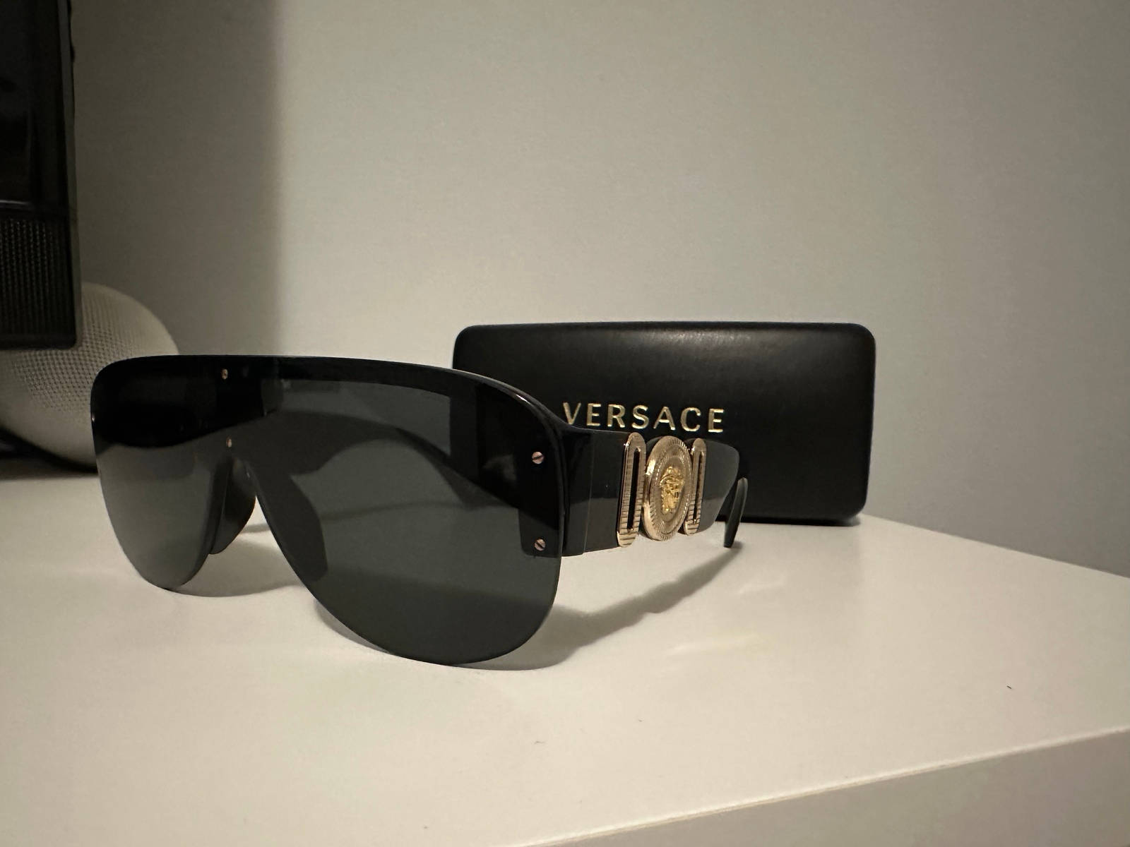 Sunglasses 桌布 壁紙 — 帶有金色美杜莎標誌的黑色Versace太陽眼鏡大膽地放在白色表面上，旁邊是其品牌眼鏡盒。