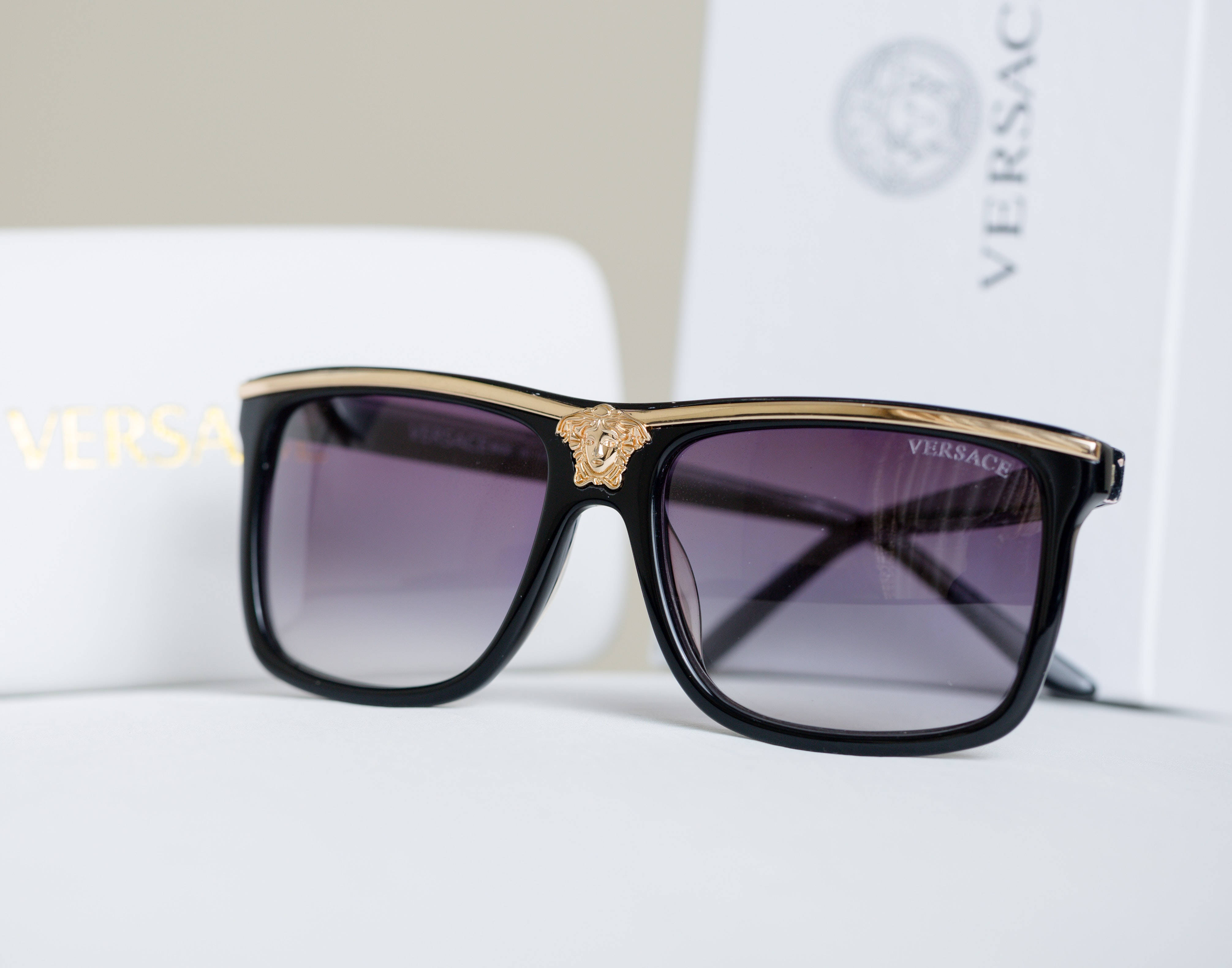 Sunglasses 桌布 壁紙 — 帶有金色美杜莎標誌的黑色Versace太陽眼鏡大膽地放在白色表面上，旁邊是品牌包裝。