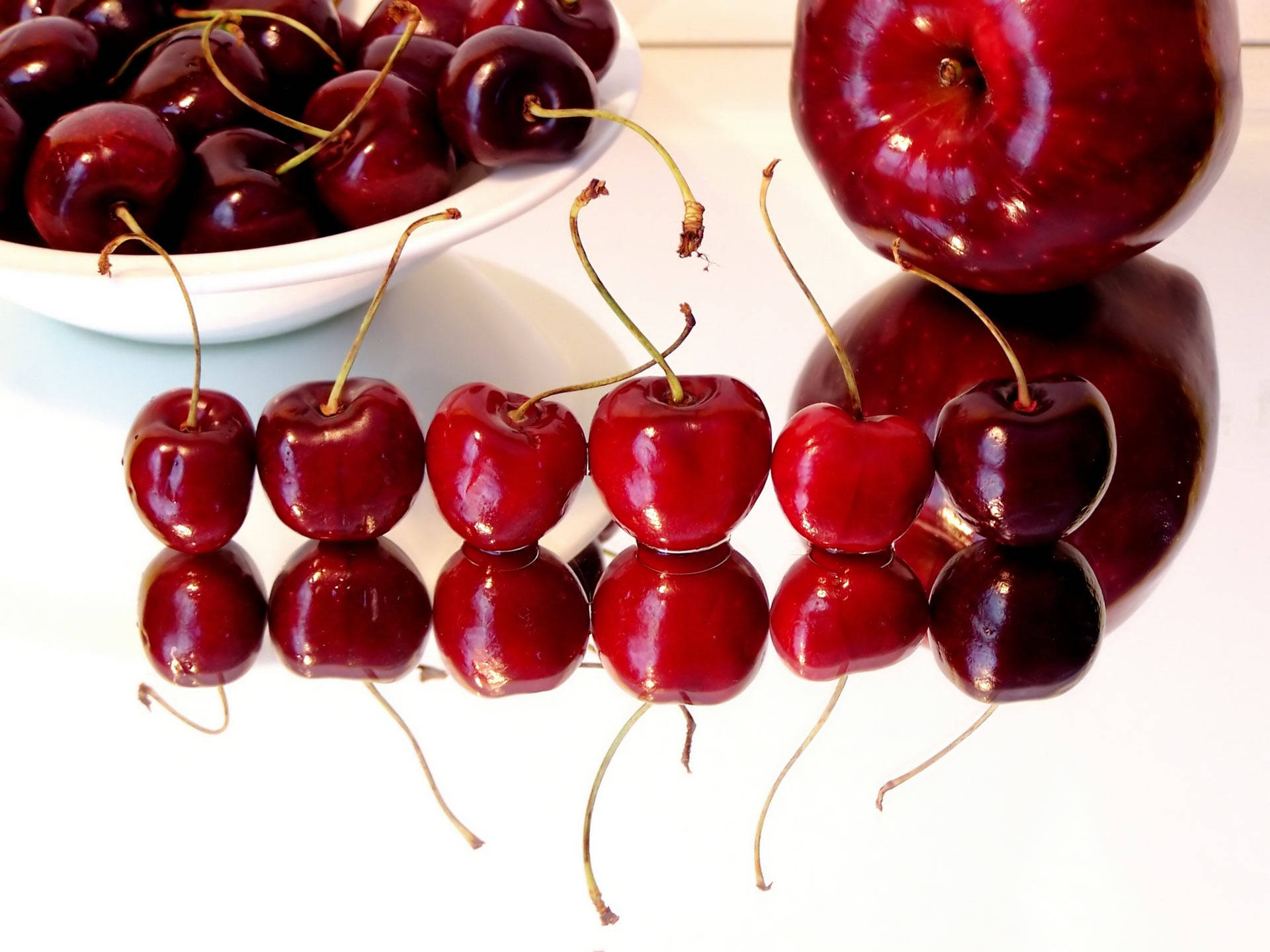 Cherry 桌布 壁紙 — 帶莖的光澤紅色櫻桃在反光白色表面上排成一排，旁邊有一碗櫻桃和一個蘋果。
