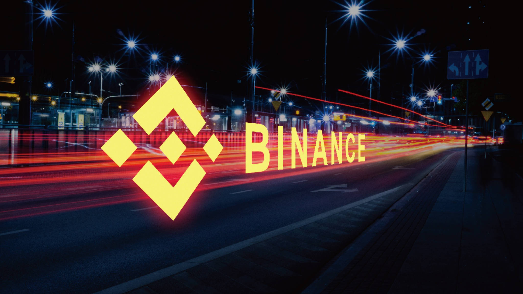 Binance 桌布 壁紙 — 幣安標誌在夜晚黑暗的城市街道上發出黃色光芒，伴隨著紅色車流燈光和星芒狀路燈。