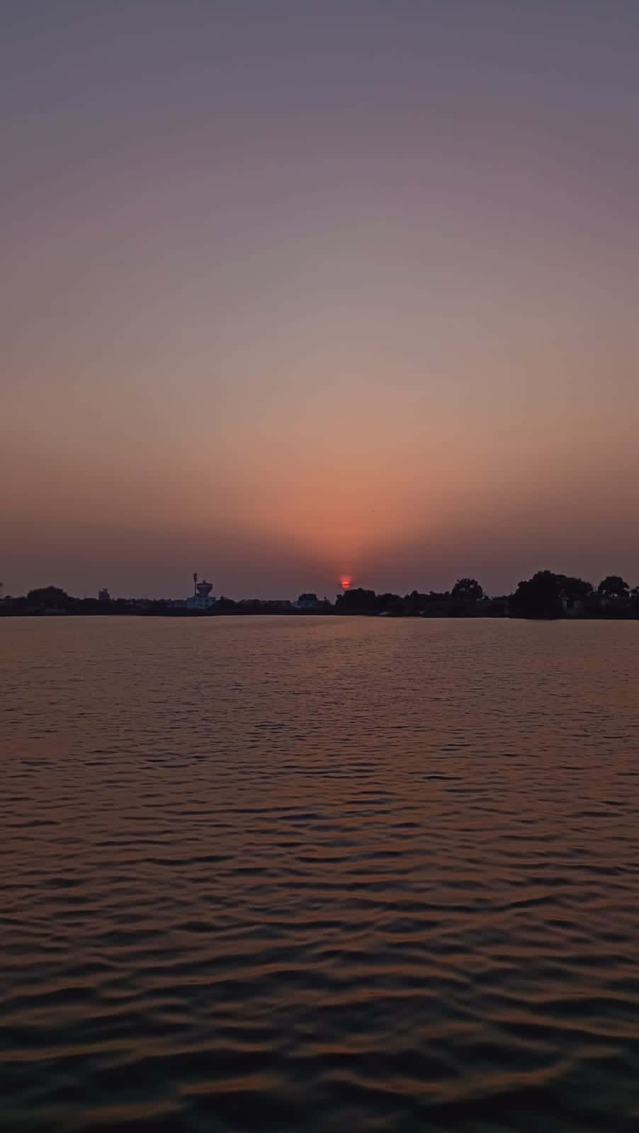 Sunset At River side 壁紙 — 平靜水面上的寧靜日落，遠方海岸線在橙色、紫色和灰色的漸變天空中形成剪影。