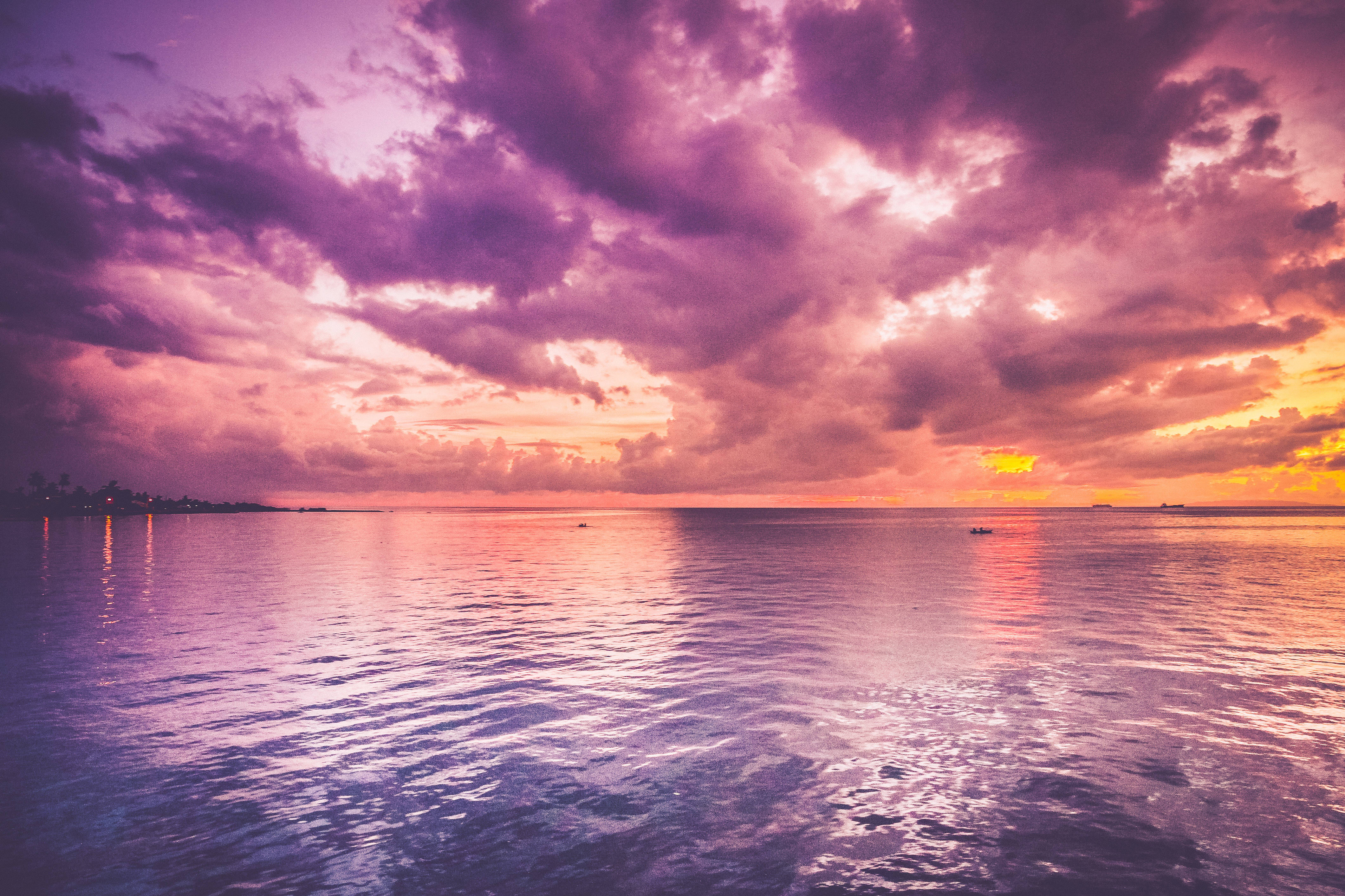 Clouds Aesthetics 桌布 壁紙 — 平靜海面上充滿活力的紫色和橙色日落，遠處的城市燈光反射在水面上。