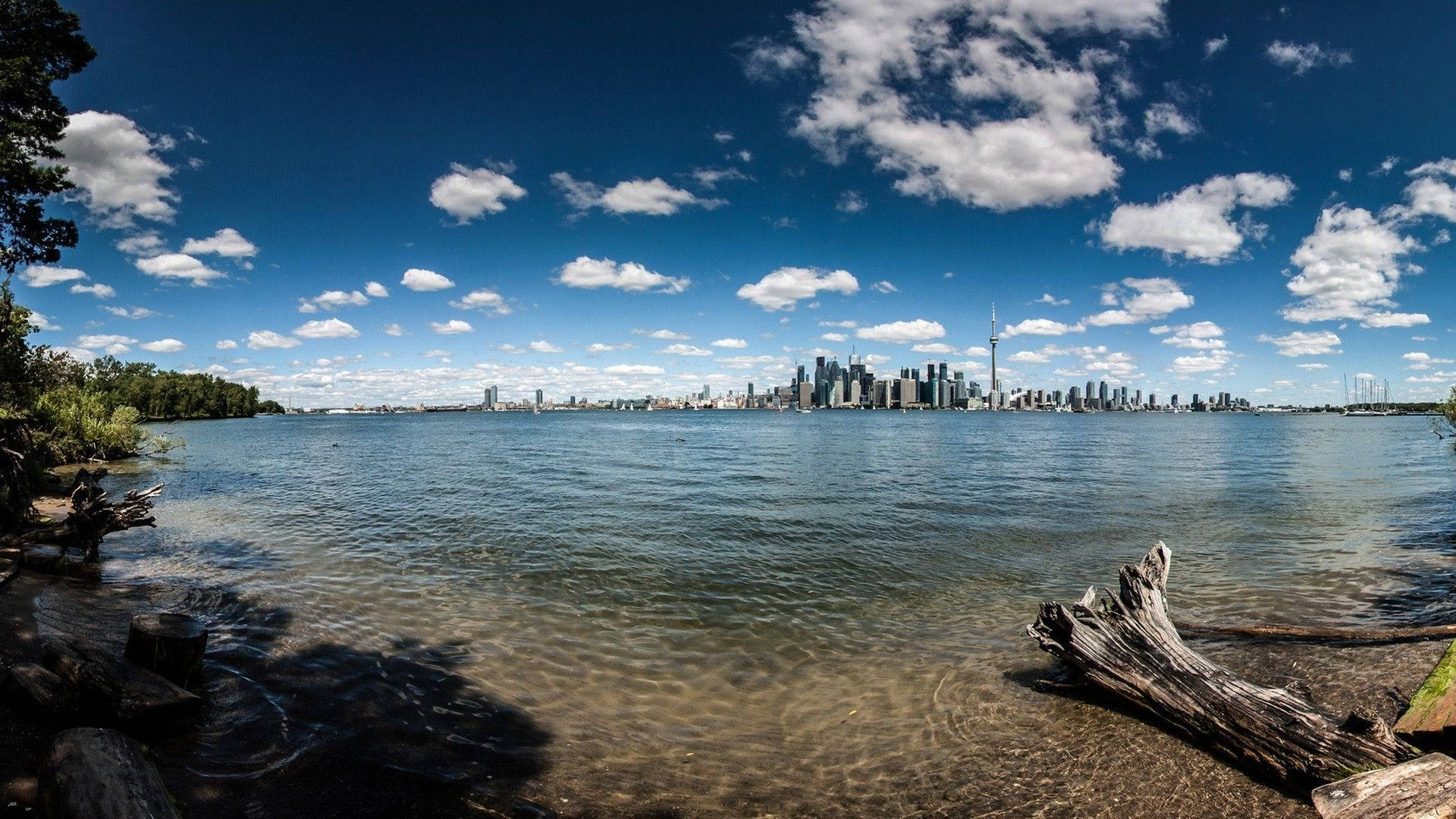 Toronto 桌布 壁紙 — 平靜湖泊對岸的現代城市天際線全景，明亮的藍天中散佈著雲朵。