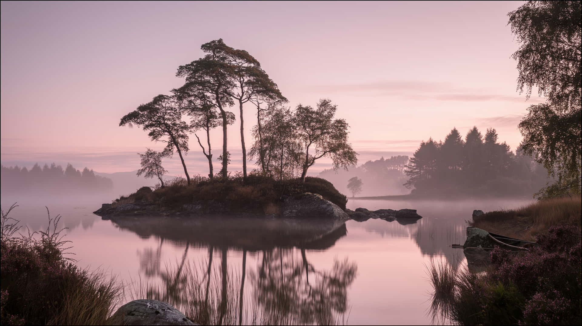Misty Lake Island at Dawn with Distant Forest Reflection 壁紙 — 平靜湖面上的霧濛濛黎明，一個小小的岩石島上長著剪影般的樹木，完美倒映在水中。