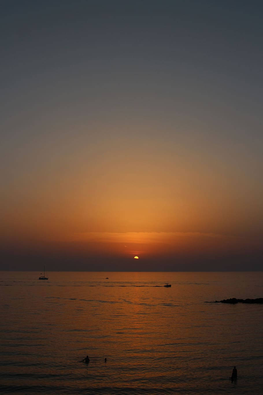 Sunset iPhone 桌布 壁紙 — 平靜的海洋上，夕陽西下，在橙色和深藍色的漸變天空下，船隻和游泳者的剪影清晰可見。