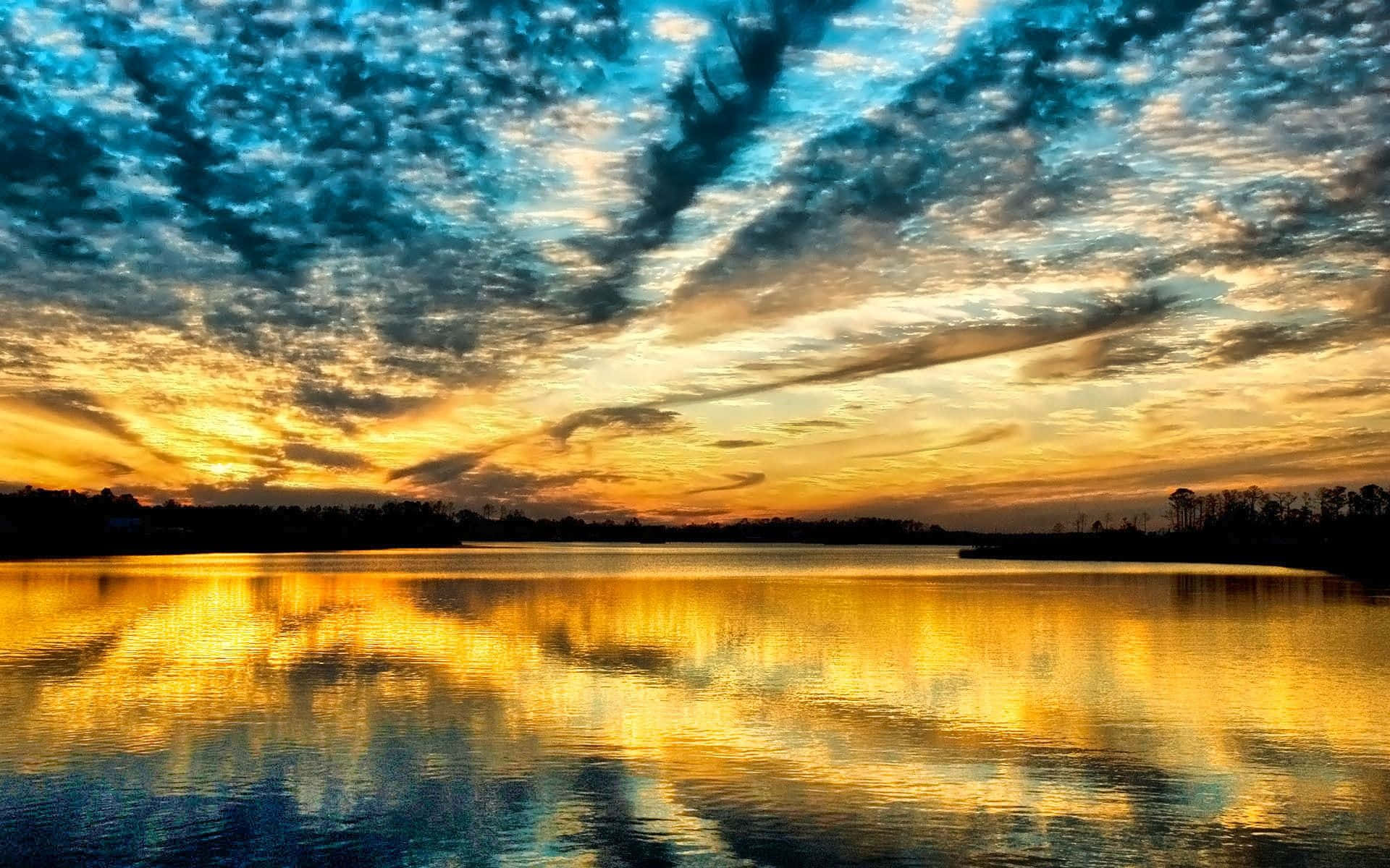 Sky Clouds 桌布 壁紙 — 平靜的湖面上寧靜的日落，金色的光線反射在水面上，上方是戲劇性的藍色雲彩。