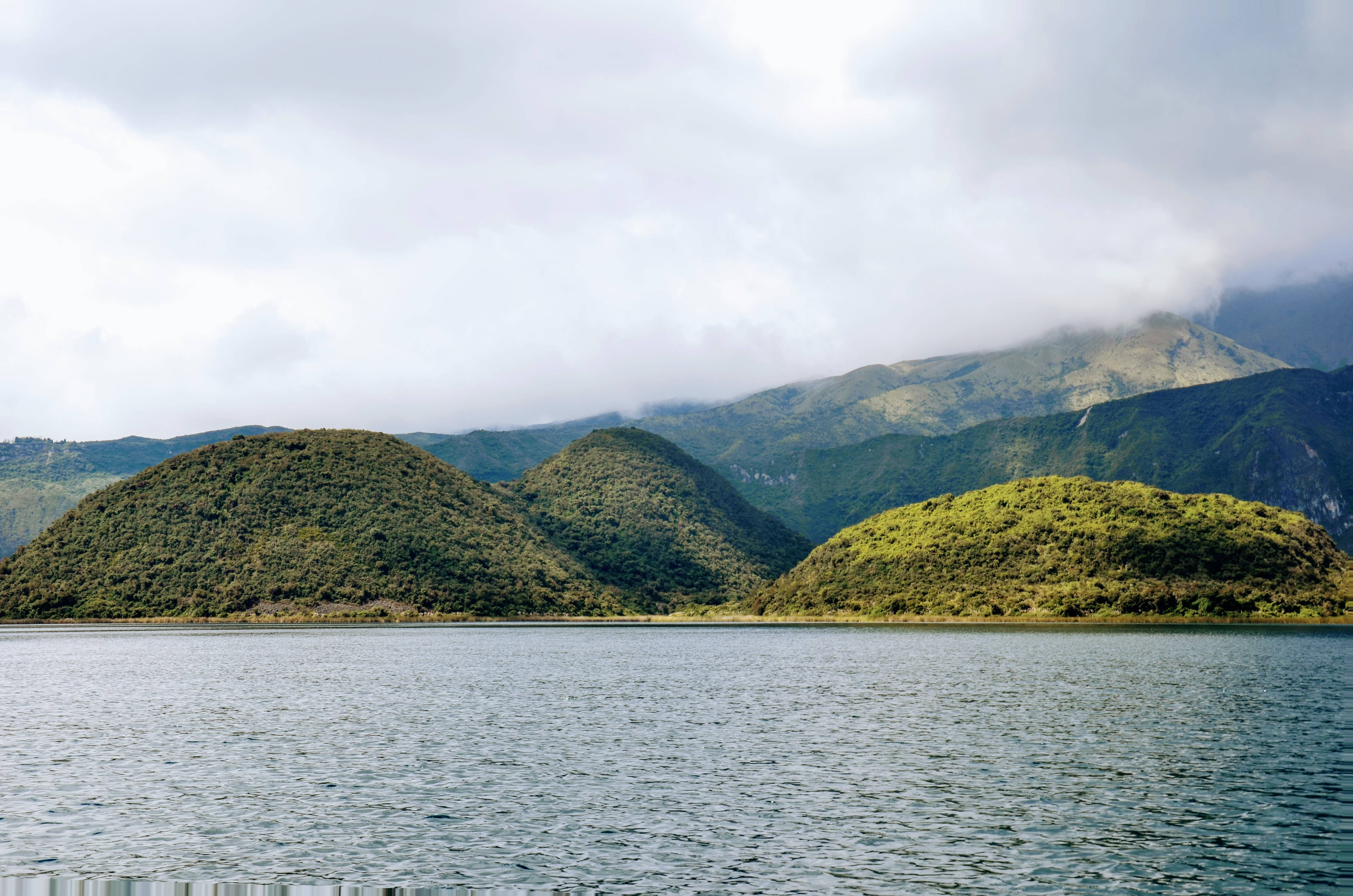 Ecuador 桌布 壁紙 — 平靜的湖面倒映著多雲的天空，起伏的綠色山丘和遠處的山脈在柔軟的雲層下升起。
