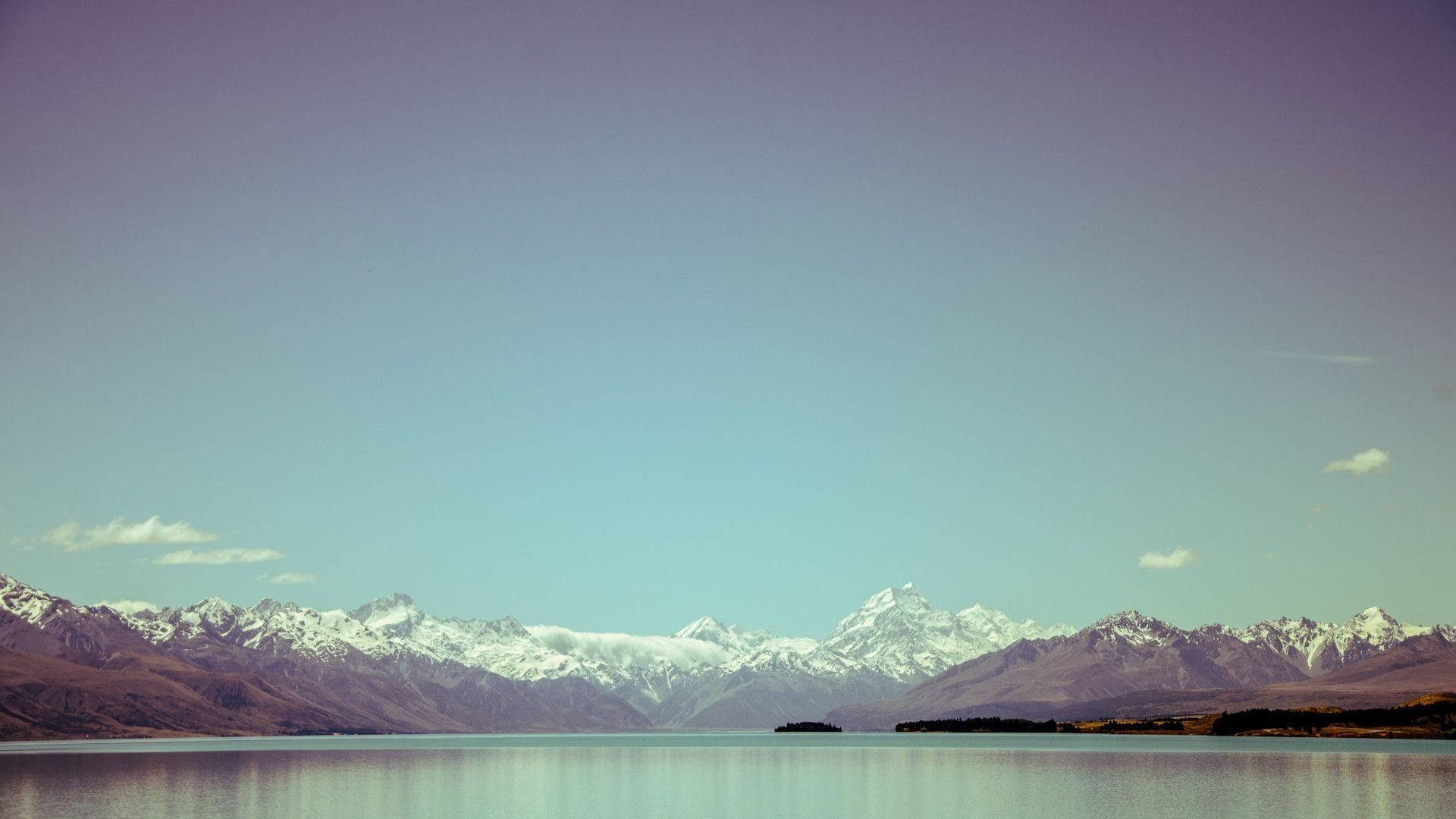 Relaxing Desktop 桌布 壁紙 — 平靜的湖面映照著白雪覆蓋的山脈，天空清澈湛藍，地平線處帶有柔和的紫色調。