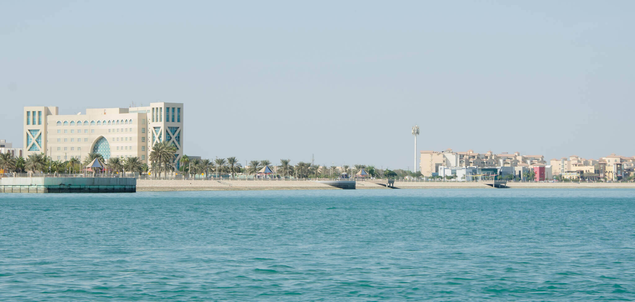 Saudi 桌布 壁紙 — 平靜的藍綠色海洋在晴朗天空下，延伸至一座擁有獨特建築和棕櫚樹的現代海岸城市景觀前。