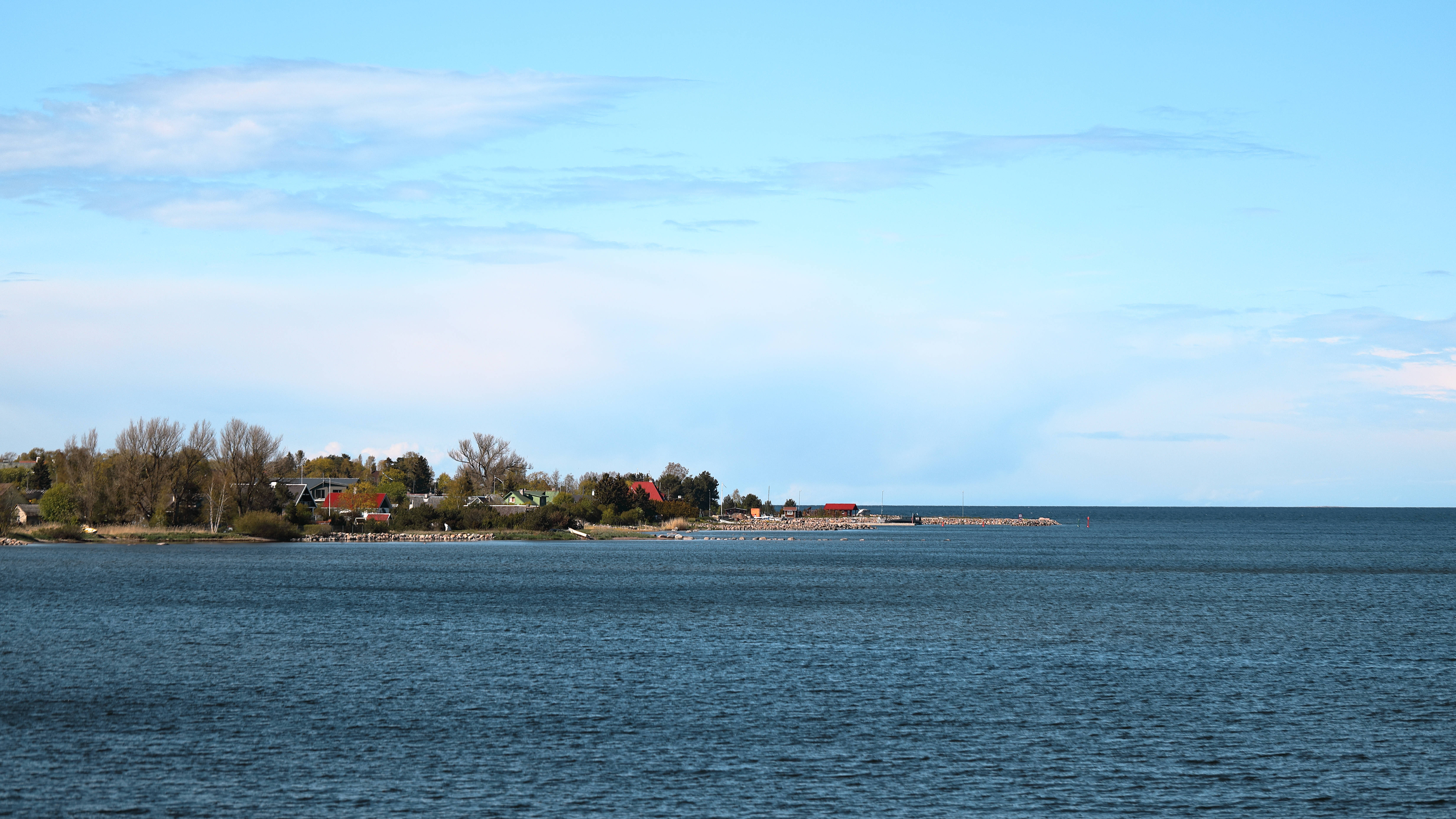 Estonia 桌布 壁紙 — 平靜的藍色湖泊延伸至遠處的湖岸，岸上有零星的房屋和樹木，天空部分多雲。