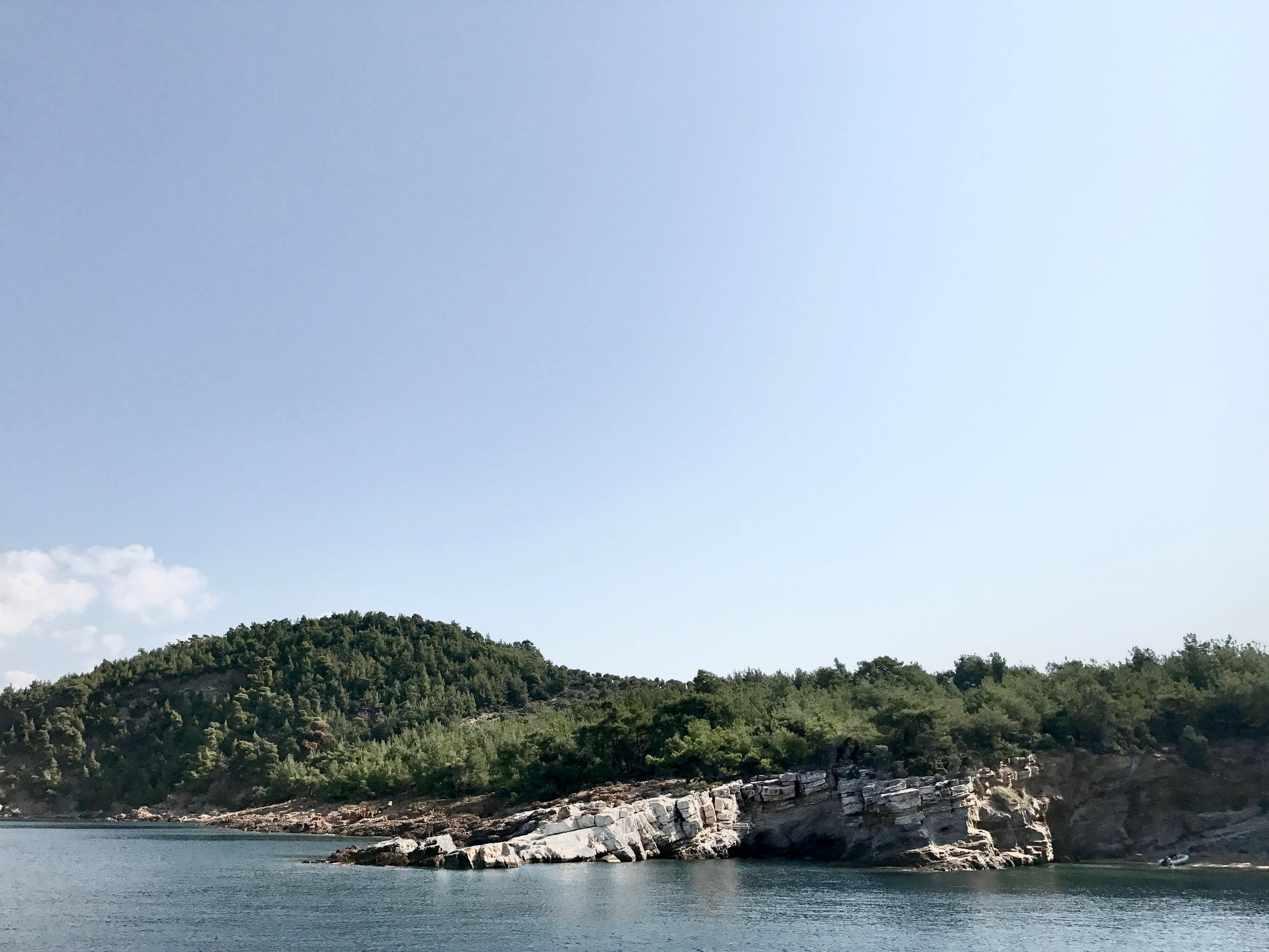 Aesthetic For Laptop 桌布 壁紙 — 平靜的藍色湖泊與岩石海岸線接壤，岸邊有茂密的綠樹，天空晴朗。