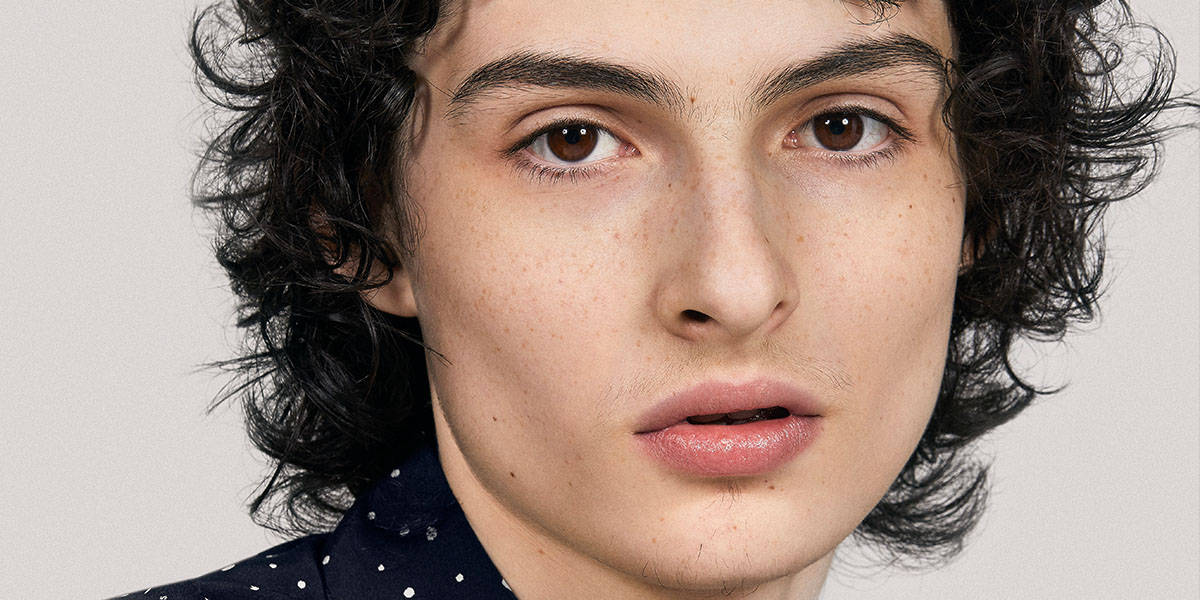 finn wolfhard 桌布 壁紙 — 年輕人的特寫肖像，深色捲髮和雀斑，背景為純淺灰色。