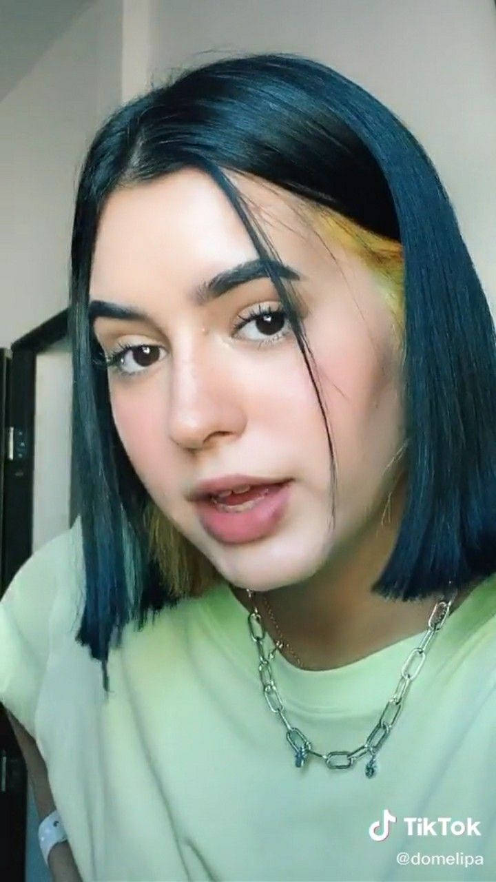 Tik Tok 桌布 壁紙 — 年輕女子特寫肖像，深藍色頭髮帶有鮮黃色條紋，佩戴銀色鏈條項鍊。