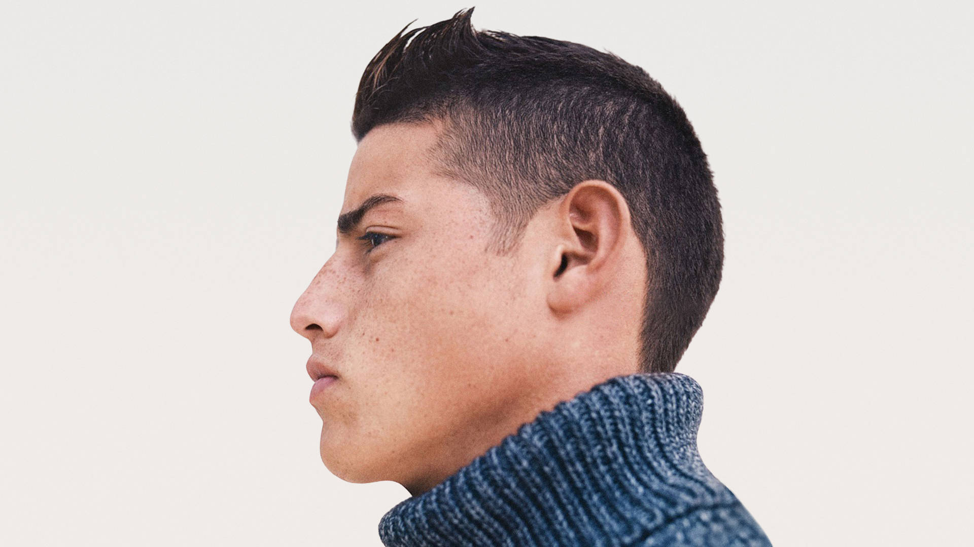 James Rodriguez 桌布 壁紙 — 年輕有雀斑的男子穿著藍色羅紋高領毛衣，以純米白色背景的特寫側面照。