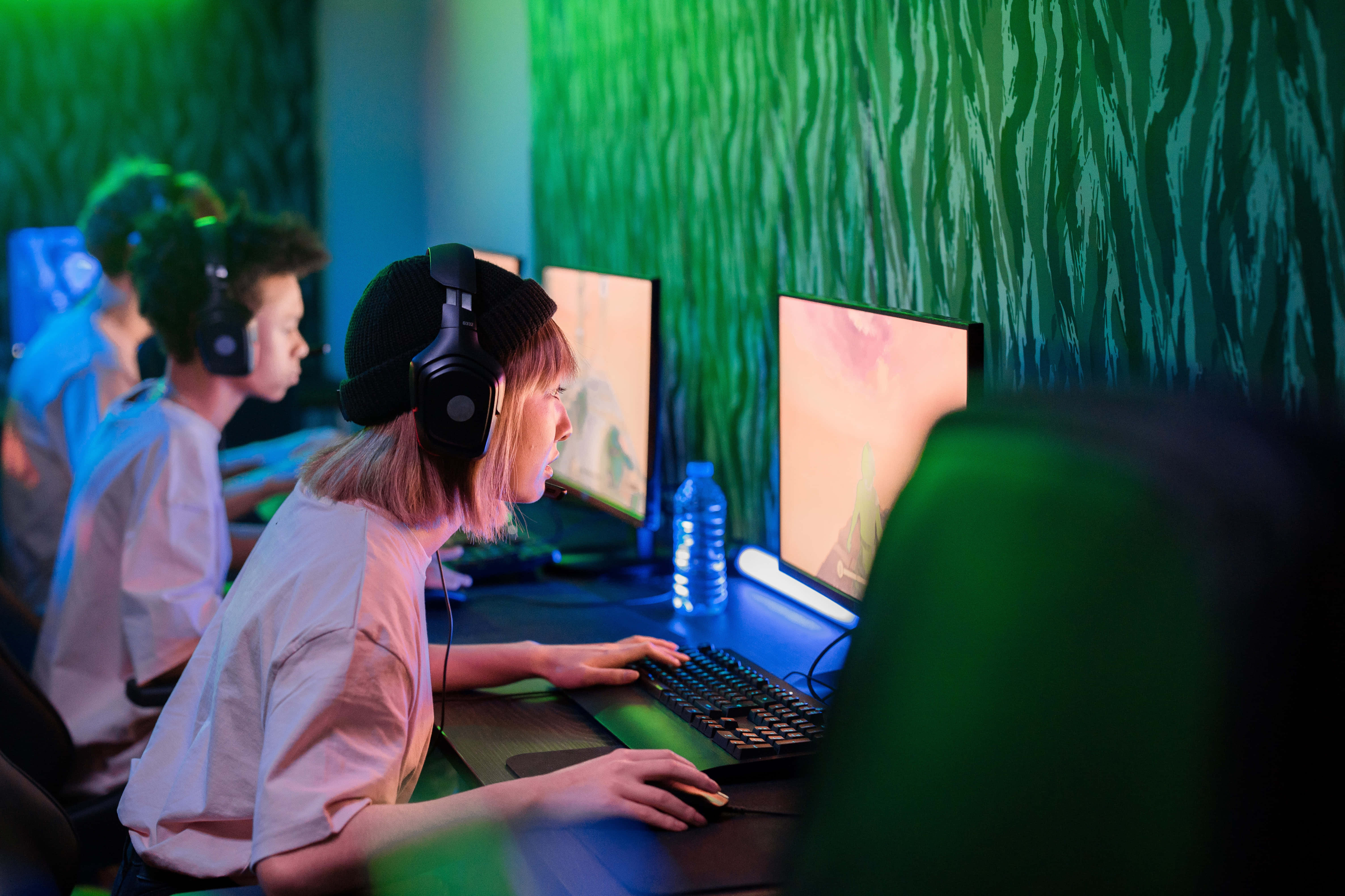Gaming Pc 桌布 壁紙 — 年輕玩家在霓虹燈照亮的房間中，專注於發光的螢幕，牆壁上有綠色圖案。