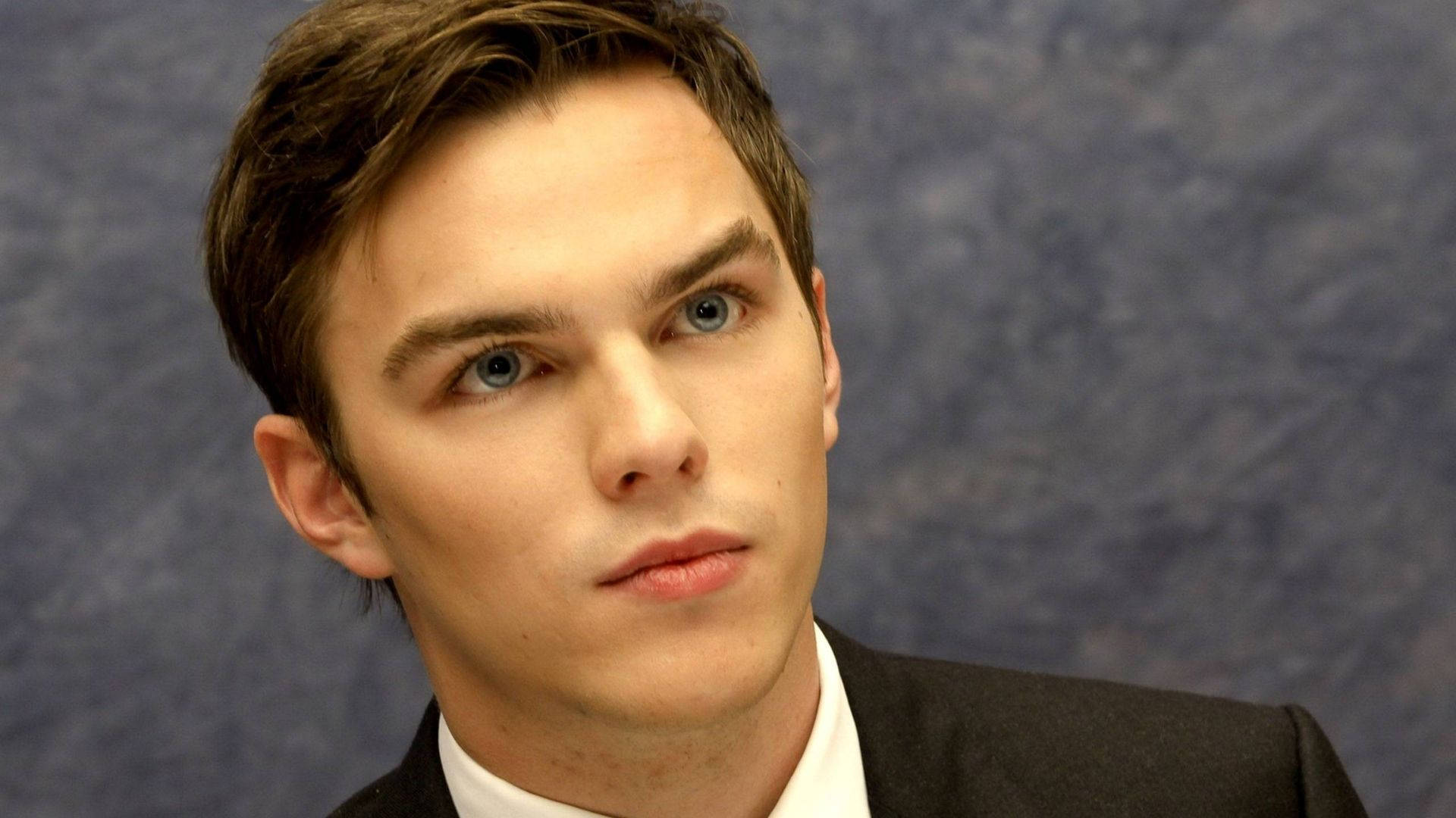 Nicholas Hoult 桌布 壁紙 — 年輕男子特寫肖像，擁有引人注目的藍色眼睛和棕色頭髮，穿著深色西裝，背景為有紋理的藍色。