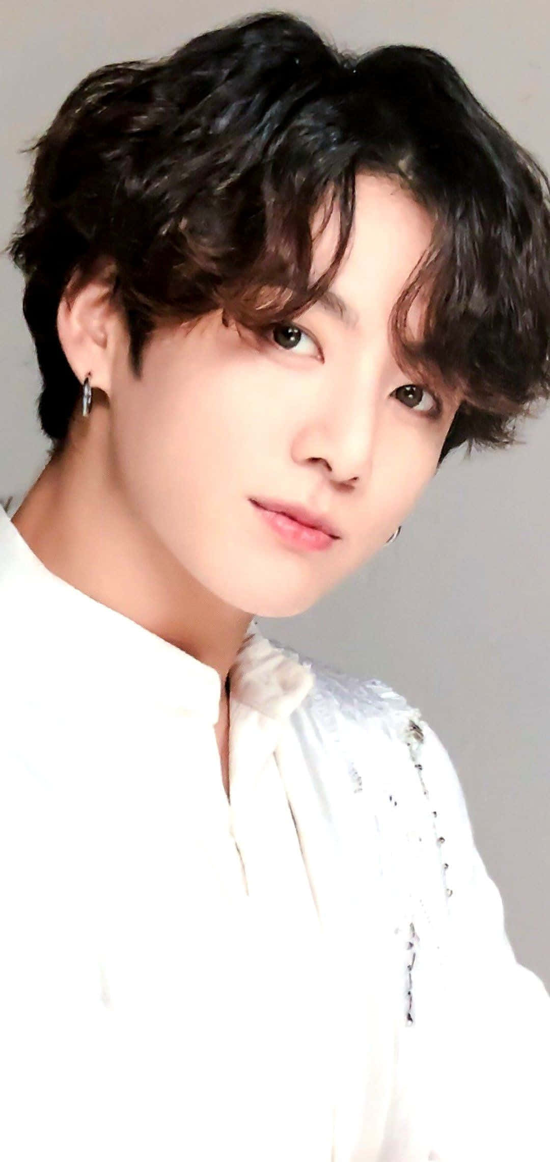 Jungkook 桌布 壁紙 — 年輕男子特寫肖像，深色捲髮，戴銀色耳環，身穿白色質感襯衫，背景為純灰色。