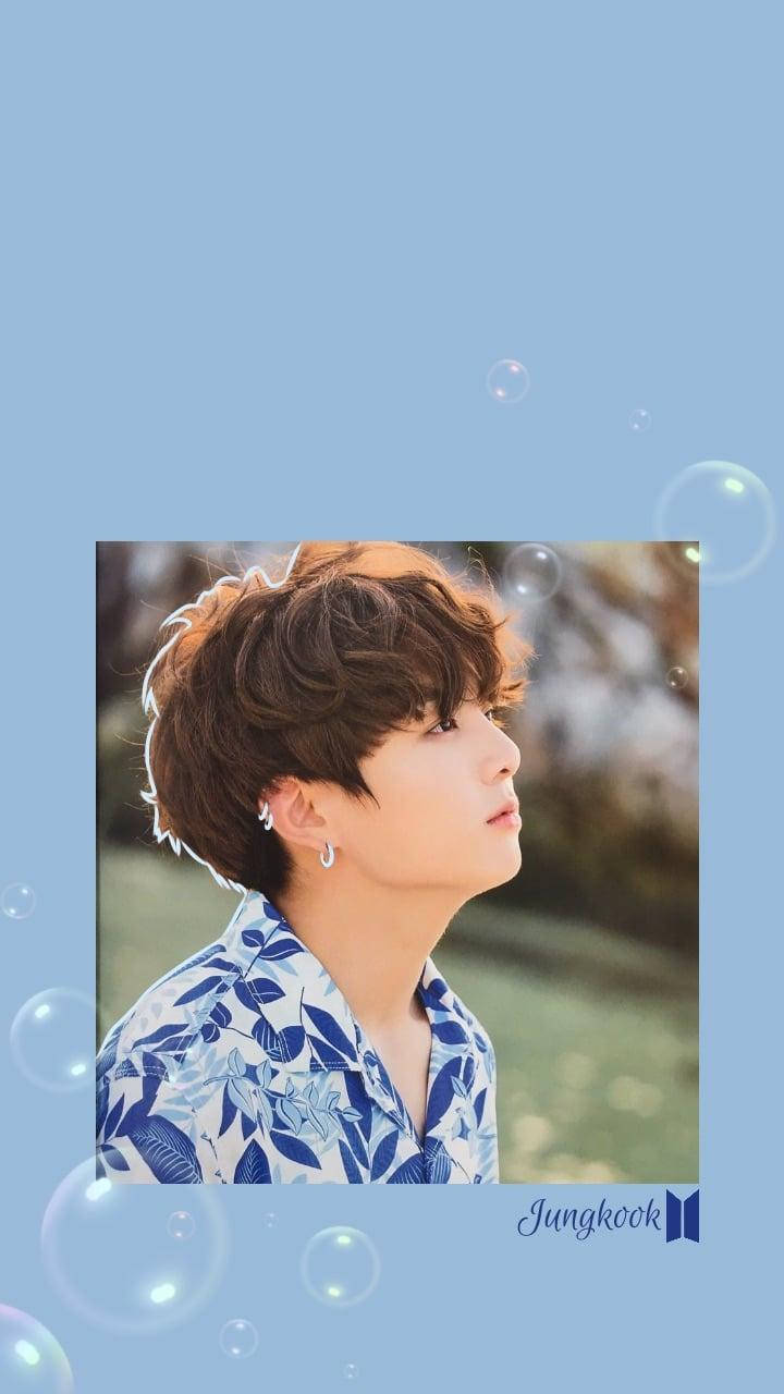 Jungkook Aesthetic 桌布 壁紙 — 年輕男子留著棕色捲髮，身穿藍色花卉襯衫，在柔和藍色背景中仰望，周圍有漂浮的氣泡。
