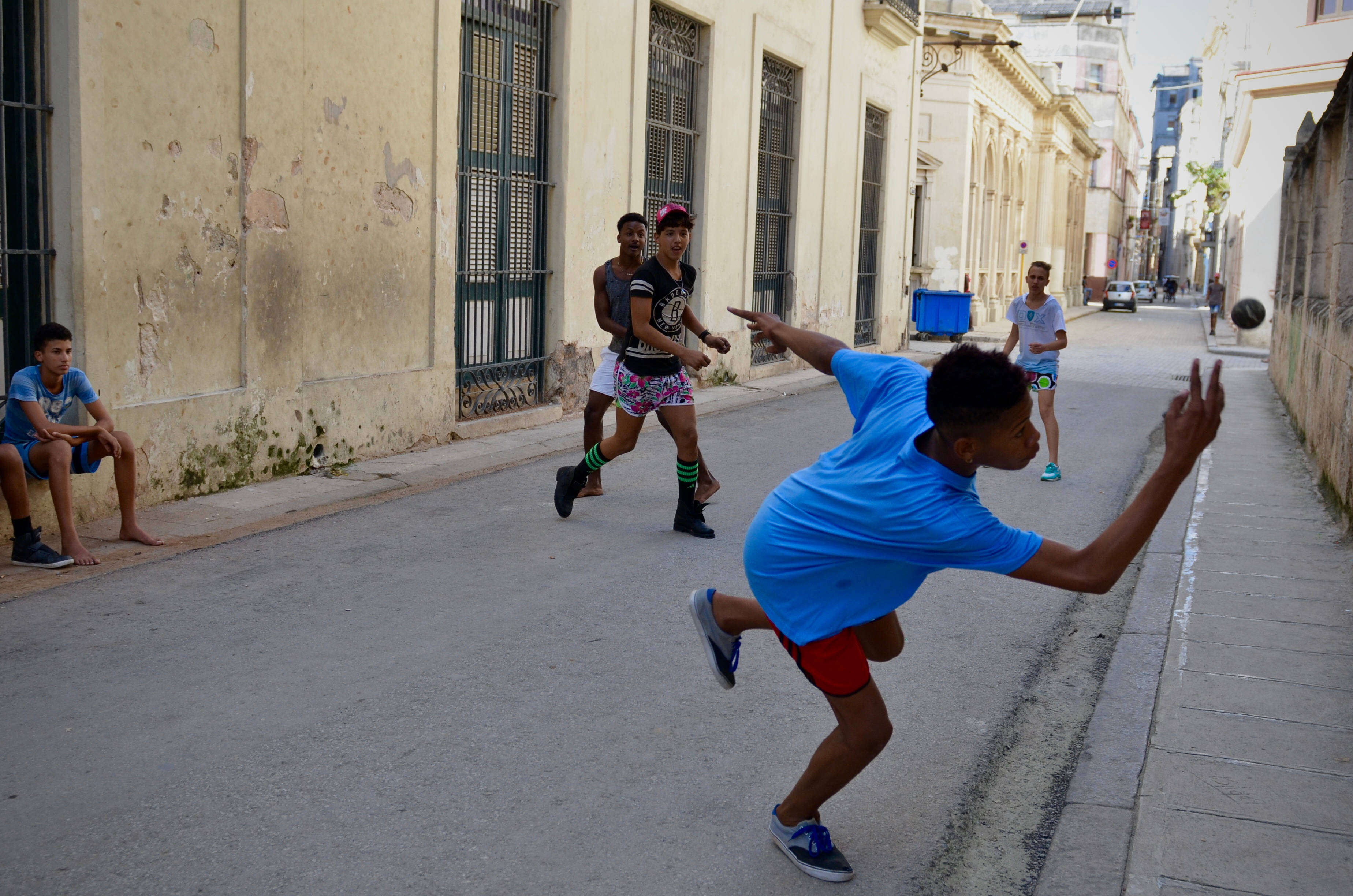 Handball 桌布 壁紙 — 年輕男孩在陽光明媚的城市小巷中充滿活力地打街頭籃球，周圍有風化的建築物和旁觀者。