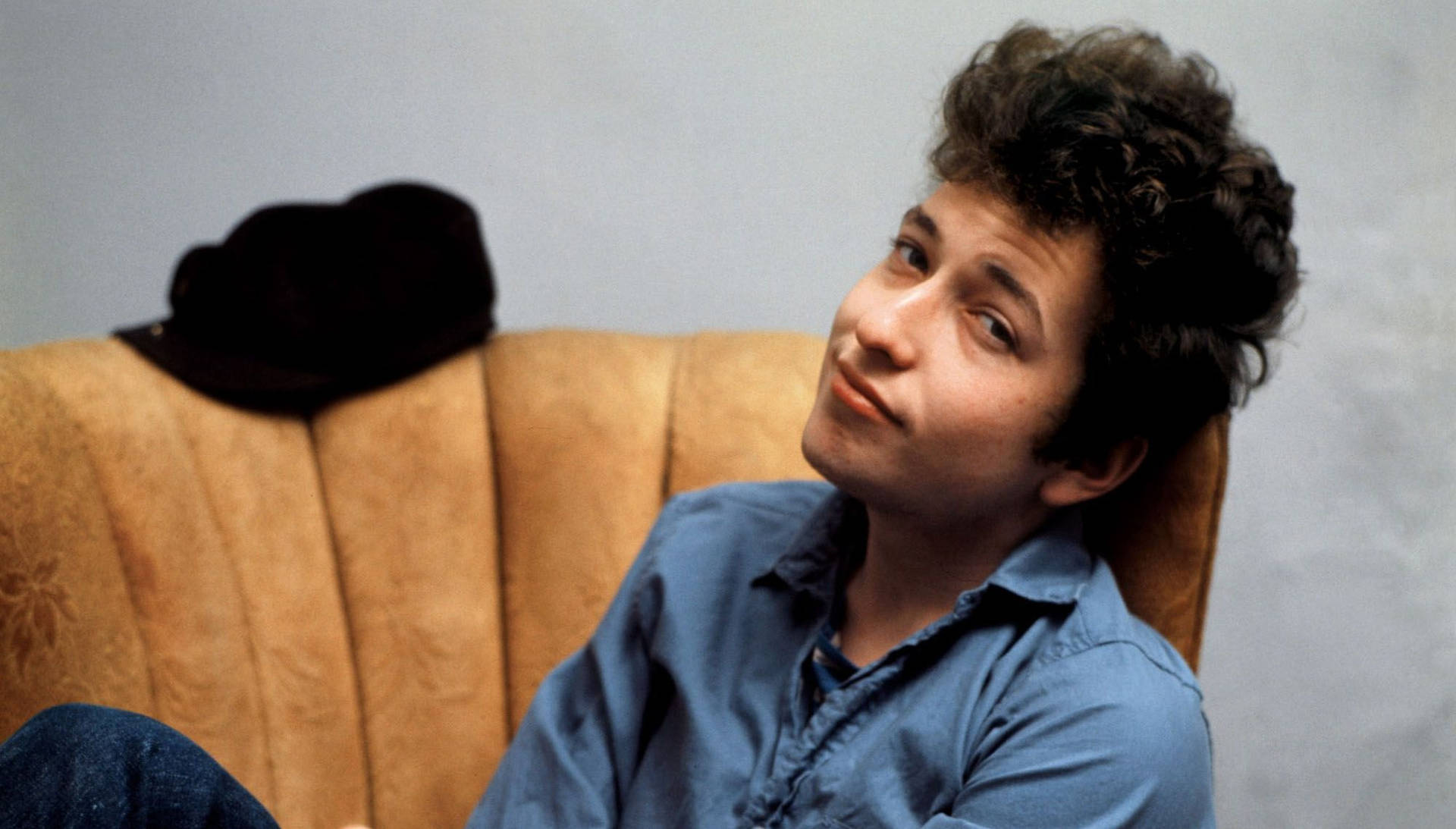  壁紙 — 年輕的Bob Dylan隨意地躺在米色沙發上，身後放著一頂黑色帽子，抬頭凝視，神情若有所思。