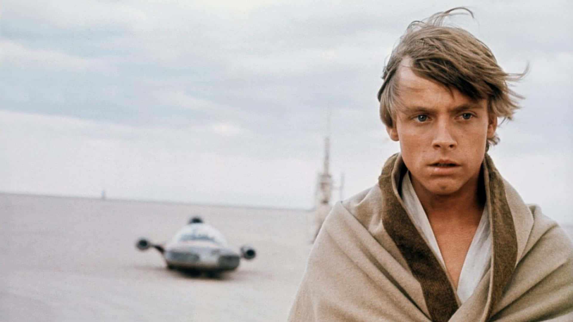 Tatooine 桌布 壁紙 — 年輕的Luke Skywalker站在荒蕪的沙漠景觀中，背景有一輛飛梭摩托車，天空多雲。