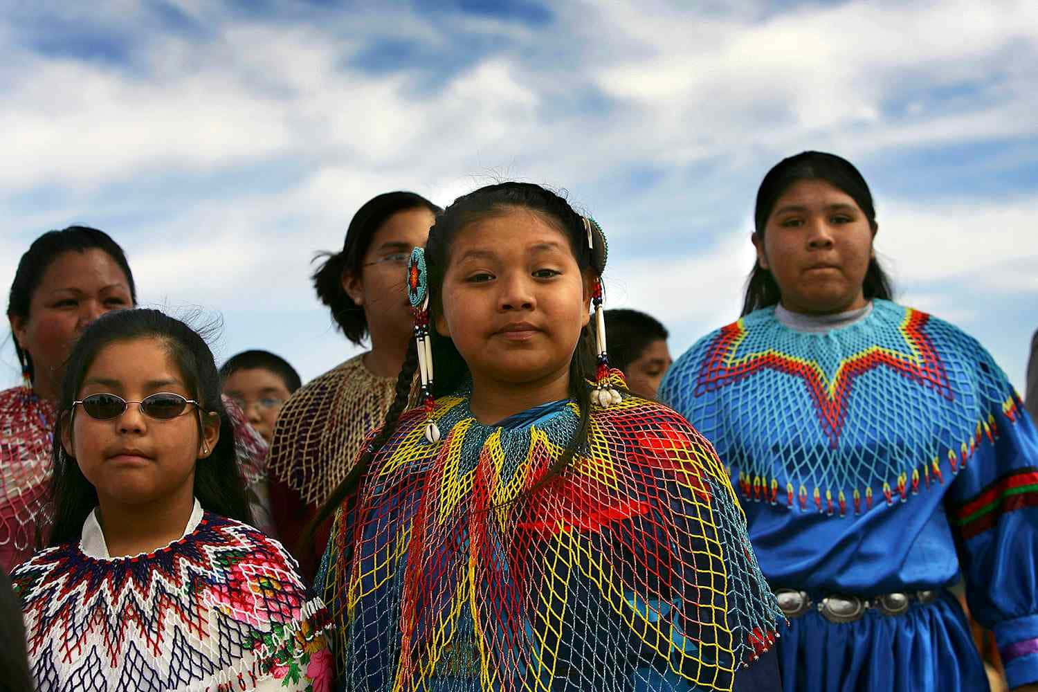 Native American 桌布 壁紙 — 年輕的原住民女孩穿著鮮豔的珠飾禮服，在部分多雲的天空下莊嚴地站立。