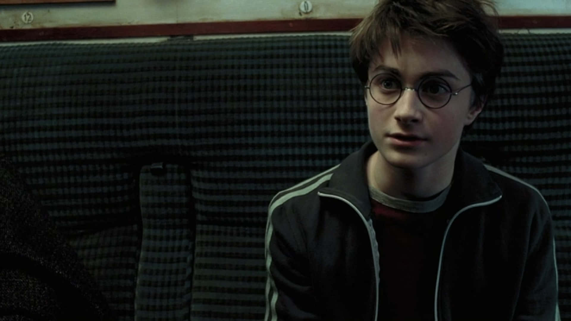 The Prisoner Of Azkaban 桌布 壁紙 — 年輕的哈利波特坐在格子火車座位上，戴著圓形眼鏡和深色外套向前看。
