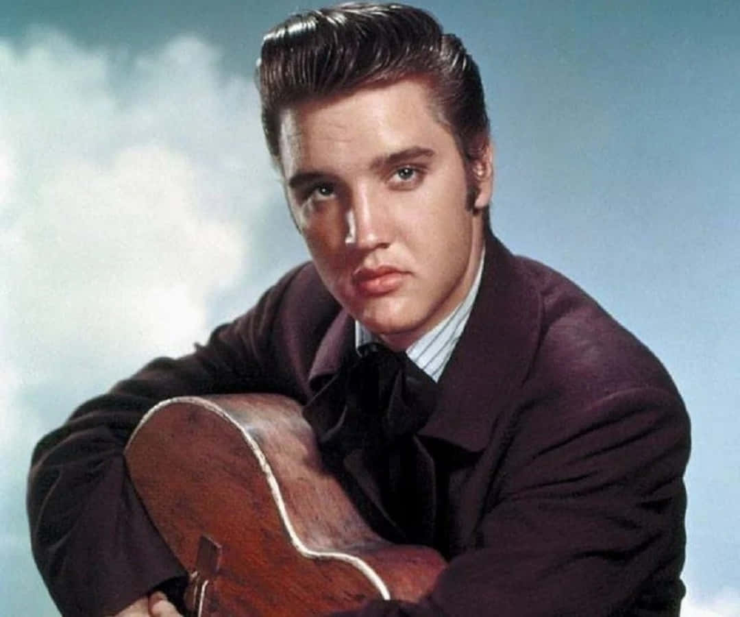 Elvis 桌布 壁紙 — 年輕的埃爾維斯·普雷斯利專注地凝視著，手持一把原聲吉他，背景是柔和的多雲天空。
