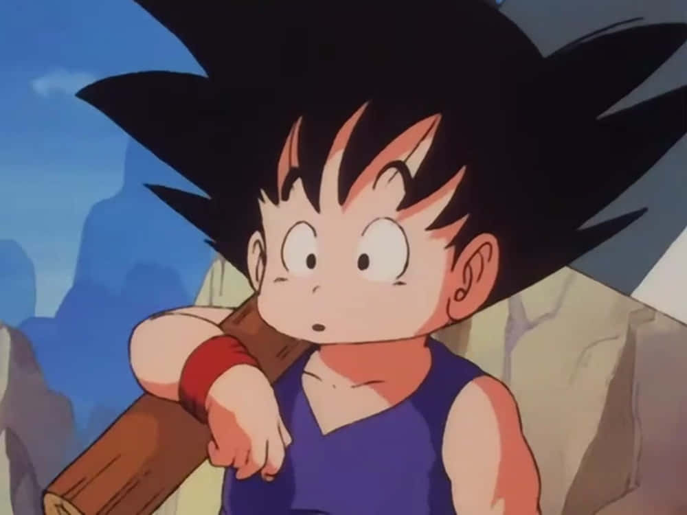 Funny Goku 桌布 壁紙 — 年輕的悟空肩上扛著一根木杖，背景是岩石山脈，天空清澈湛藍。
