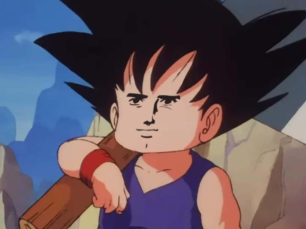 Funny Goku 桌布 壁紙 — 年輕的悟空自信地站著，肩上扛著一根木杖，背景是岩石峭壁，頭頂是晴朗的藍天。