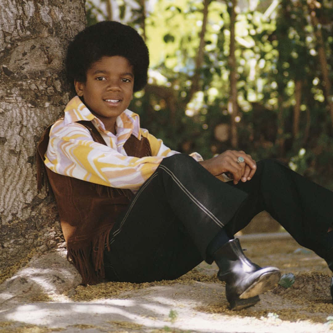 Young Michael Jackson 桌布 壁紙 — 年輕的非洲式髮型男孩微笑著靠著樹幹坐在陽光斑駁的森林裡，穿著圖案襯衫和流蘇背心。