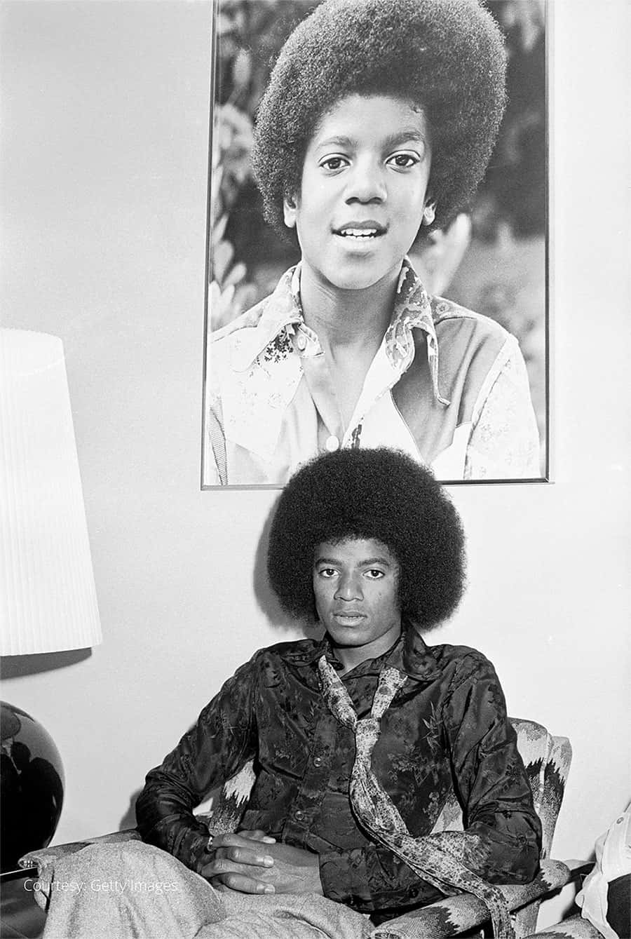 Young Michael Jackson 桌布 壁紙 — 年輕的麥可·傑克森坐在椅子上，下方掛著自己的肖像，兩者均帶有標誌性的非洲爆炸頭和圖案襯衫，在單色室內環境中。