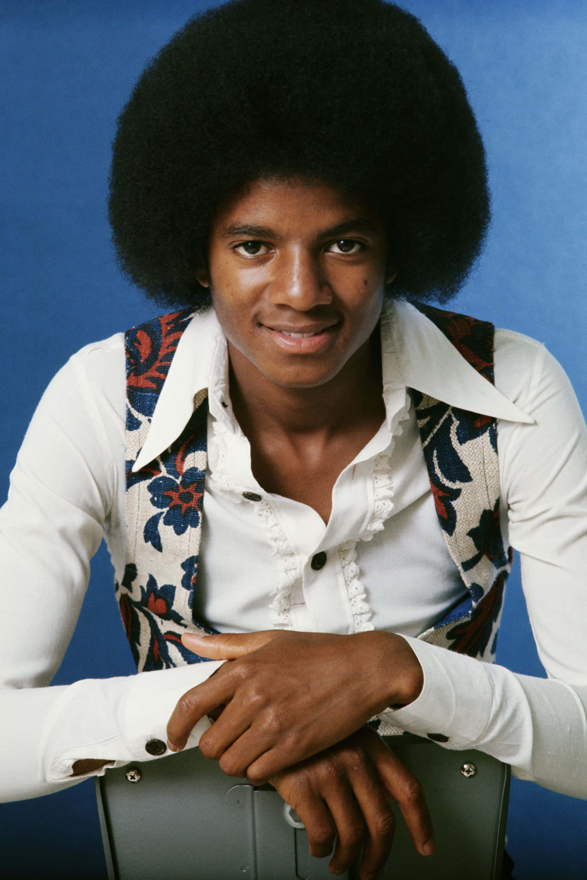 Young Michael Jackson 桌布 壁紙 — 年輕的麥可傑克森留著大爆炸頭，身穿花卉背心，在純藍色攝影棚背景下溫暖微笑。