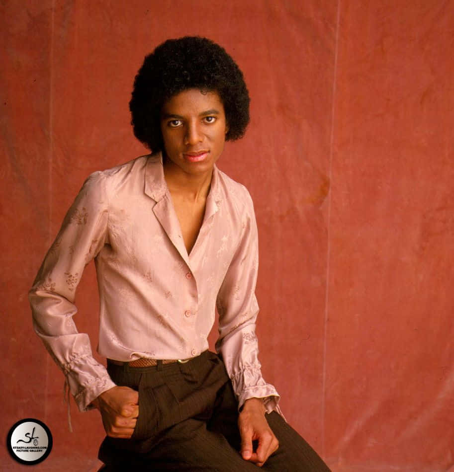 Young Michael Jackson 桌布 壁紙 — 年輕的麥可傑克森自信地坐在有紋理的陶土背景前，身穿粉紅色圖案襯衫和深色褲子。