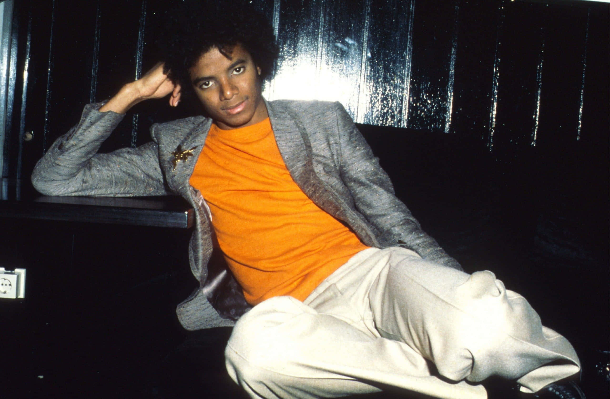 Young Michael Jackson 桌布 壁紙 — 年輕的麥可傑克森自信地穿著灰色西裝外套和橙色高領毛衣，靠在深色鑲板牆上，燈光效果戲劇性。