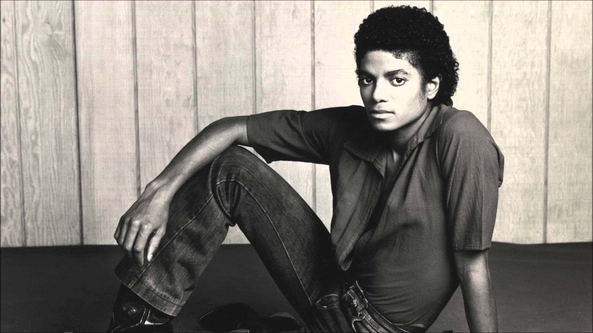 Young Michael Jackson 桌布 壁紙 — 年輕的麥可傑克森隨意地靠著木牆坐著，一幅引人注目的黑白肖像。