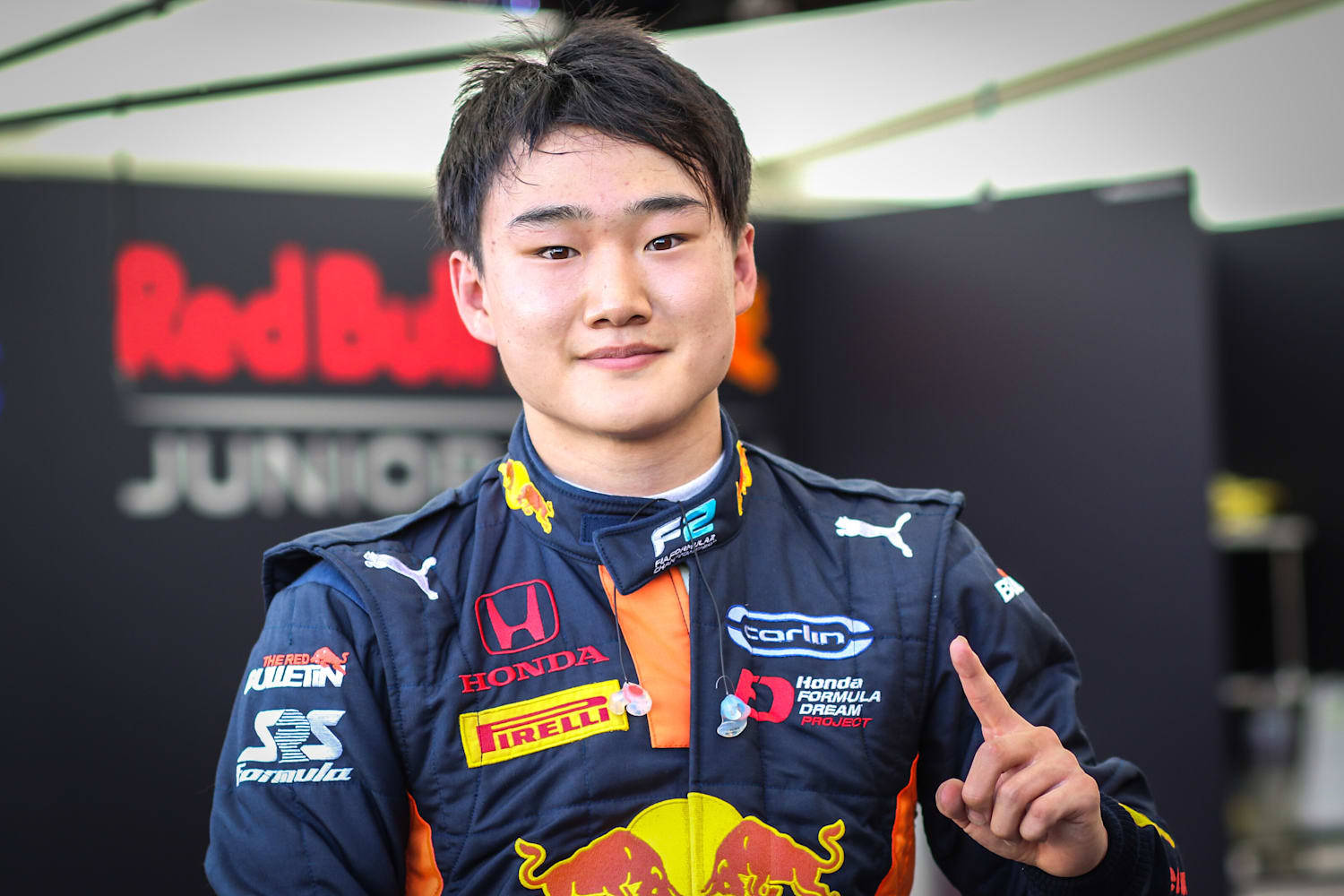 Racing 桌布 壁紙 — 年輕賽車手身穿 Red Bull Junior 裝備，自信地指向鏡頭，穿著亮橙色和黑色的賽車服。