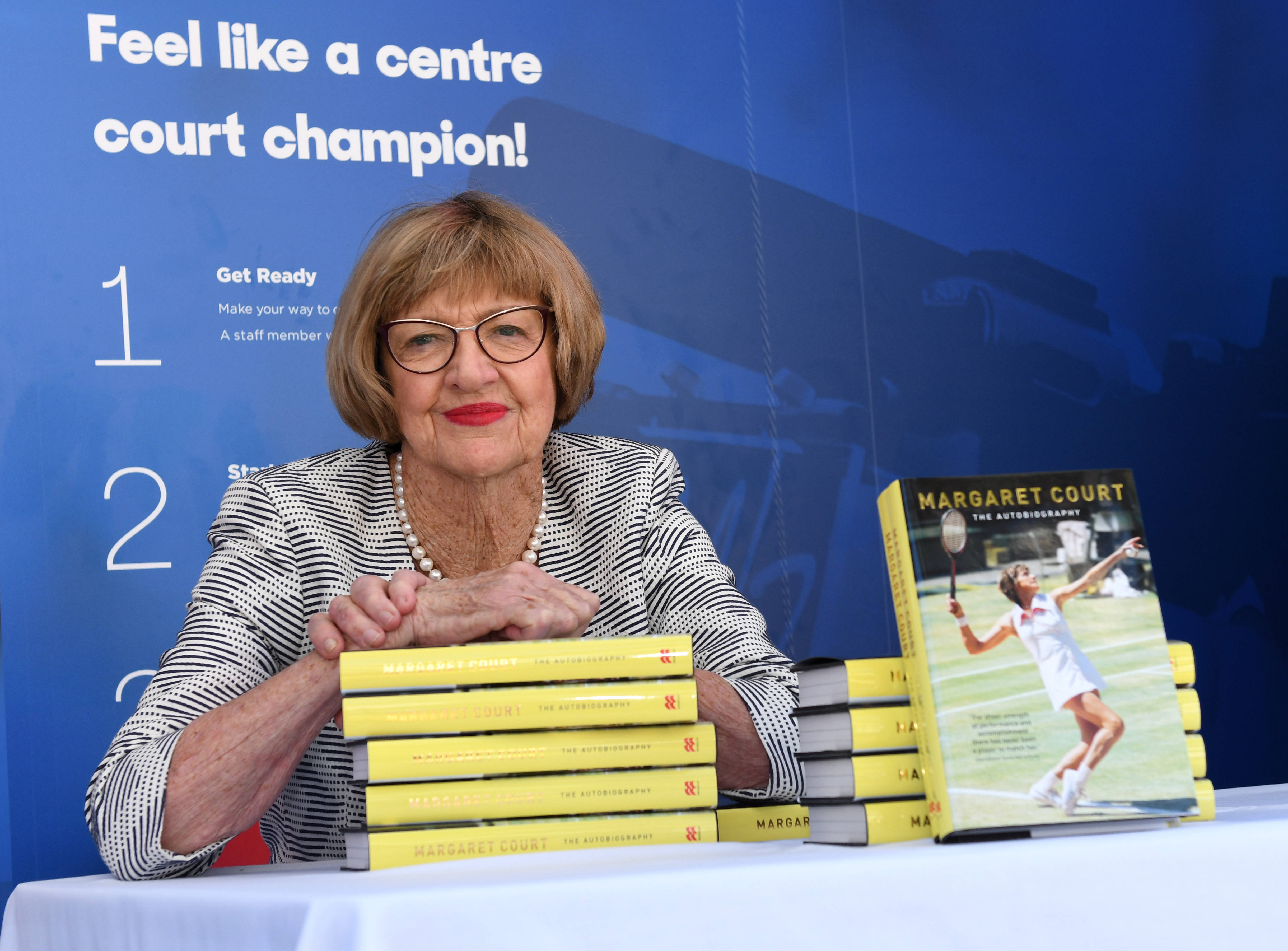 網球 桌布 壁紙 — 年長的網球傳奇人物 Margaret Court 在宣傳活動中坐在一堆黃色自傳書後面，露出溫暖的笑容。