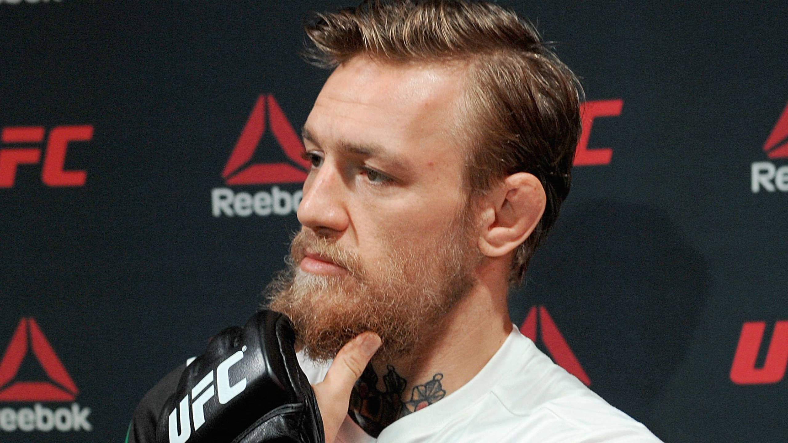 Mcgregor 桌布 壁紙 — 康納·麥格雷戈身穿白色襯衫和UFC手套，在帶有紅色UFC和Reebok標誌的深色背景下擺出嚴肅的表情。