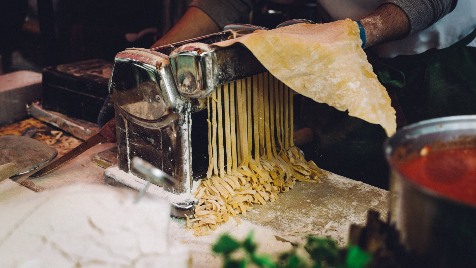 Pasta 桌布 壁紙 — 廚師將麵團放入復古義大利麵機，擠出新鮮金黃色的麵條，落在撒滿麵粉的檯面上，旁邊有一鍋紅醬。
