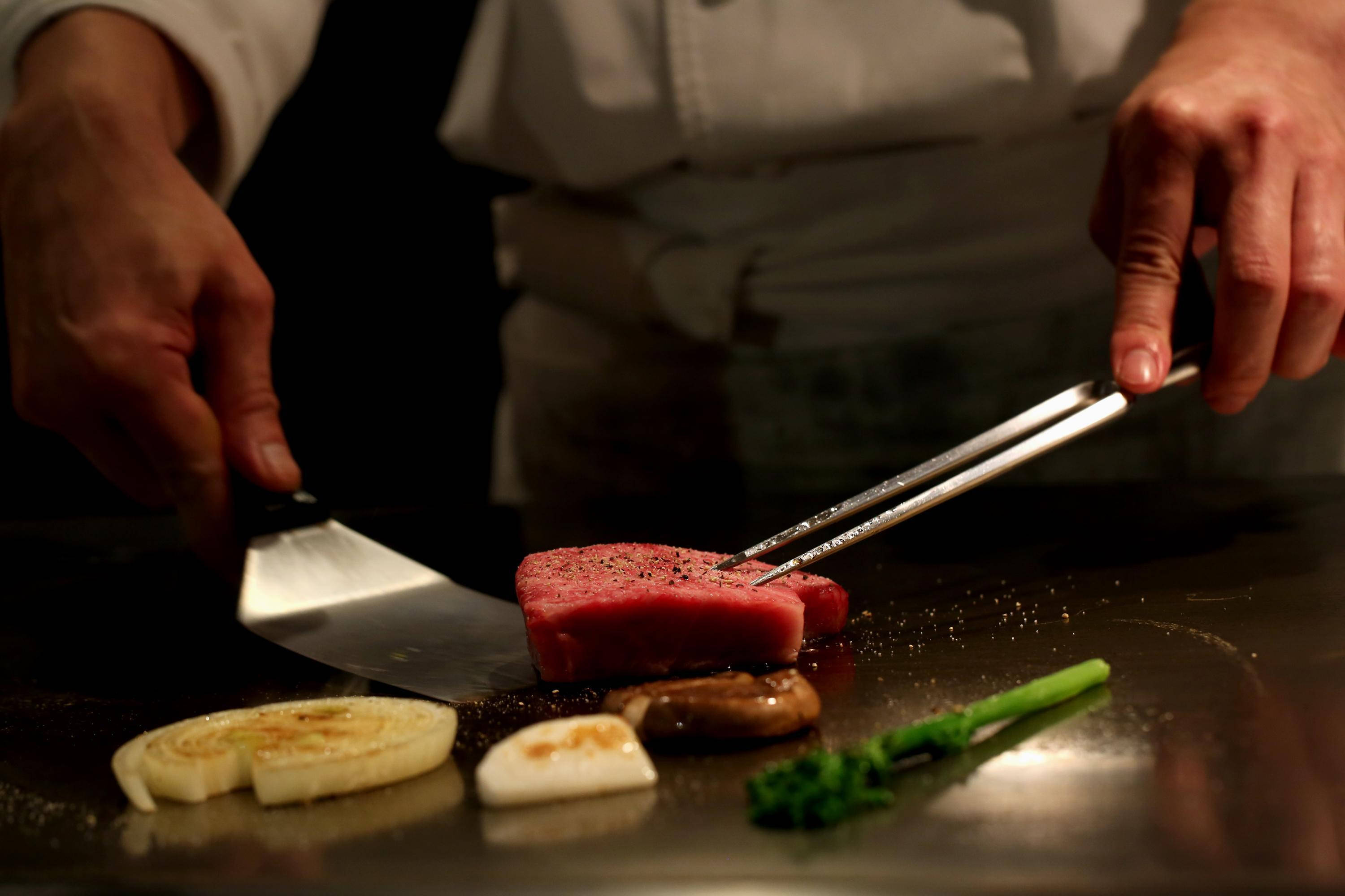 meat 桌布 壁紙 — 廚師用夾子和鍋鏟在熱騰騰的鐵板燒烤盤上煎一塊鮮紅色的鮪魚排，周圍有滋滋作響的洋蔥和青花菜。