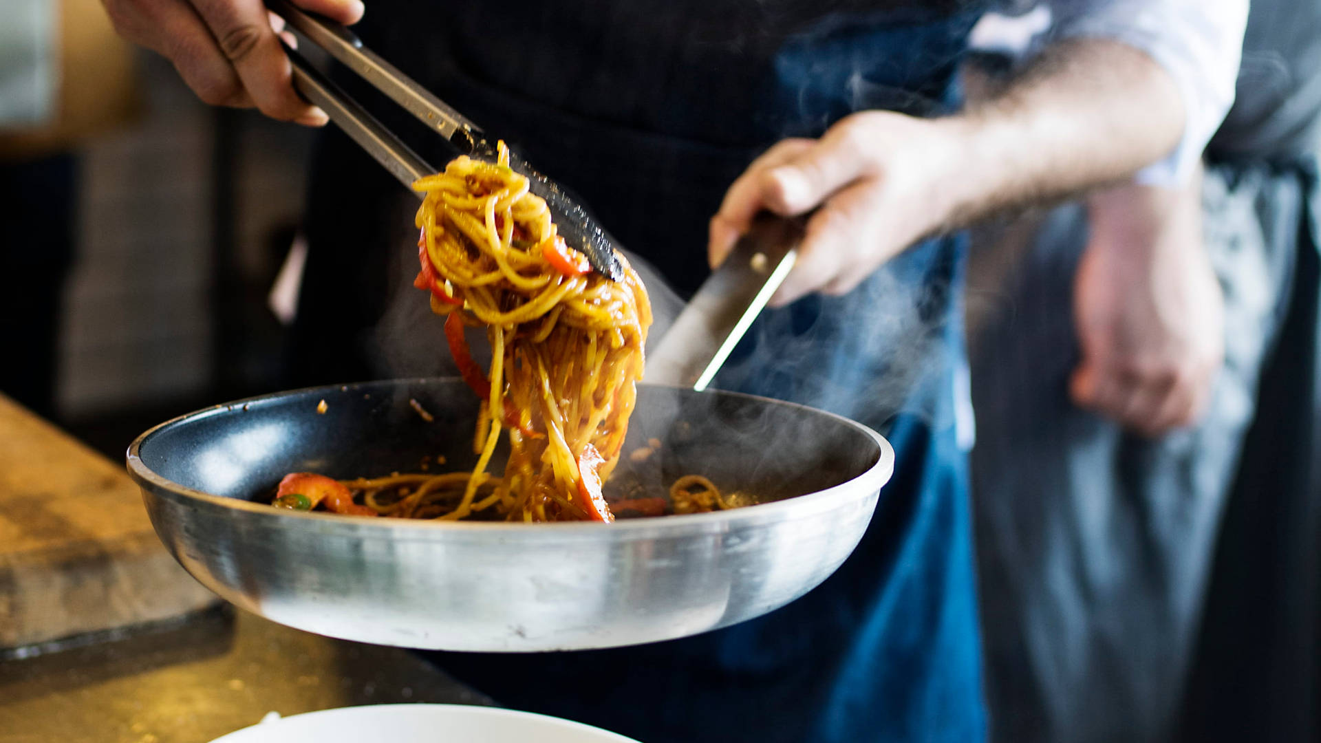 Pasta 桌布 壁紙 — 廚師用夾子從熱鍋中提起冒煙的義大利麵，廚房裡一片繁忙。