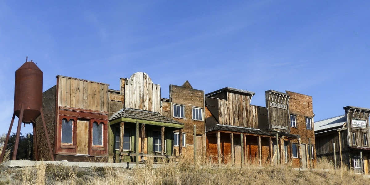 Old Western 桌布 壁紙 — 廢棄的西部小鎮木建築在晴朗的藍天下風化，前景有乾草。
