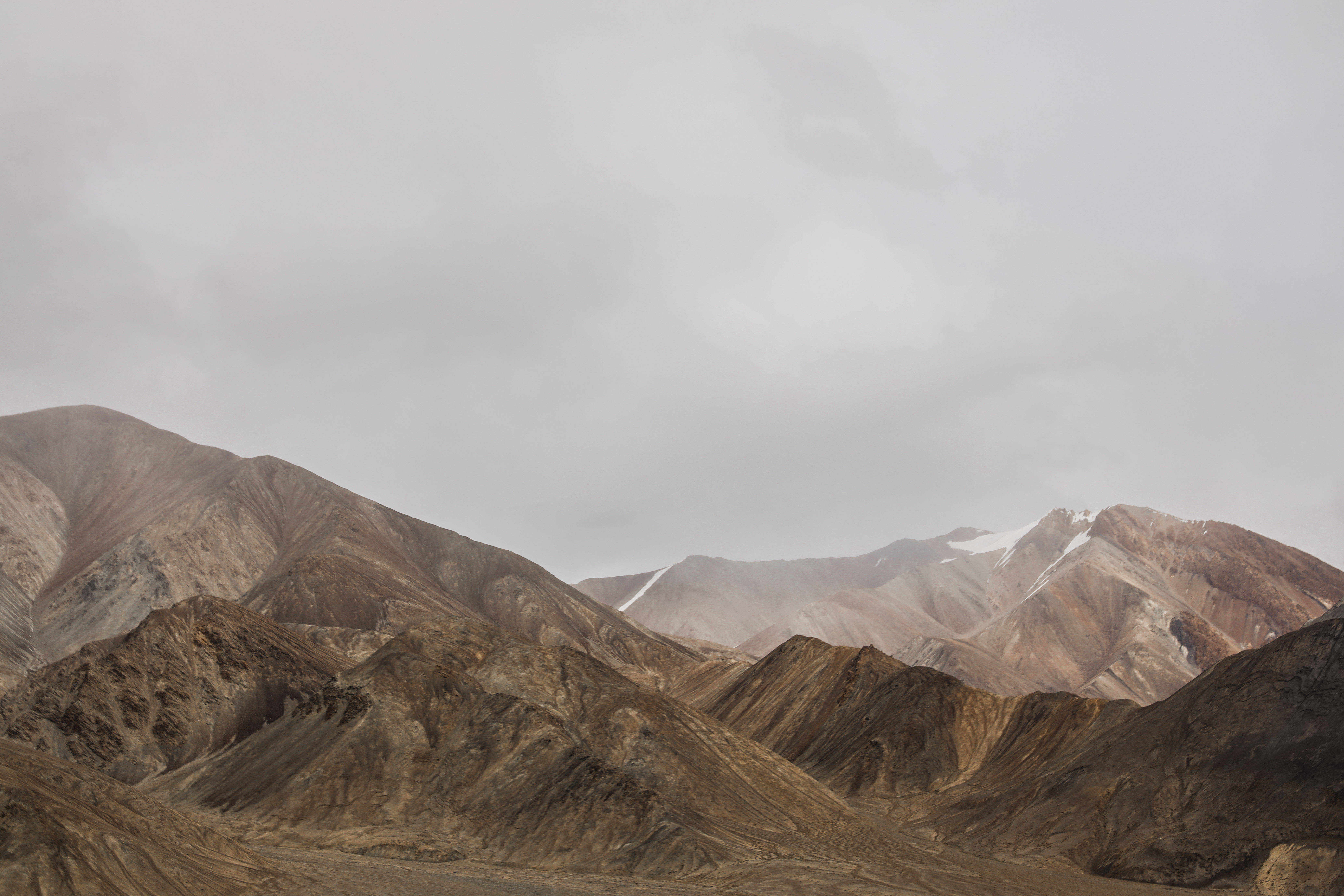 Tajikistan 桌布 壁紙 — 廣闊乾旱的山脈，遠處有白雪覆蓋的山峰，天空陰沉密佈。