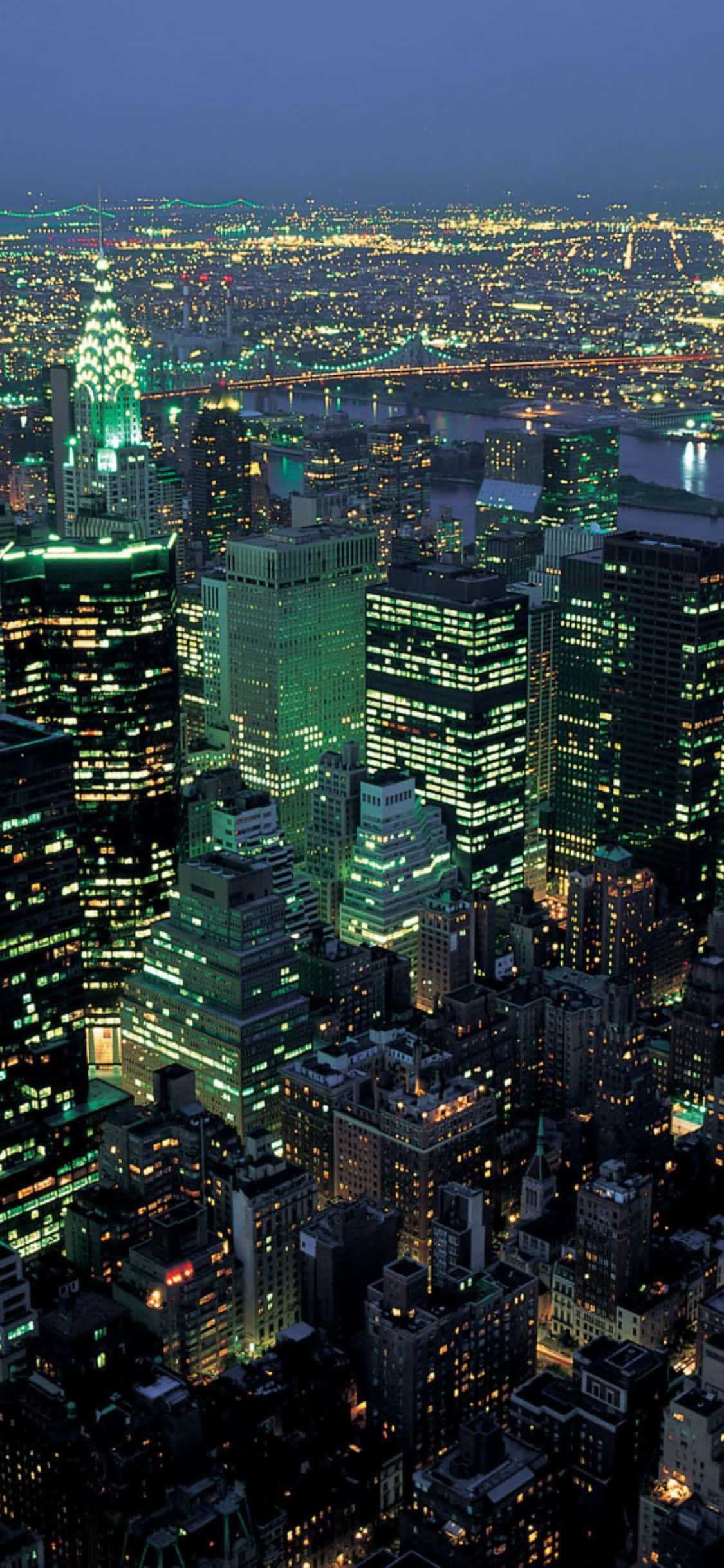 iPhone X New York 桌布 壁紙 — 廣闊城市夜景的鳥瞰圖，摩天大樓和橋樑在深藍色天空的映襯下閃閃發光。